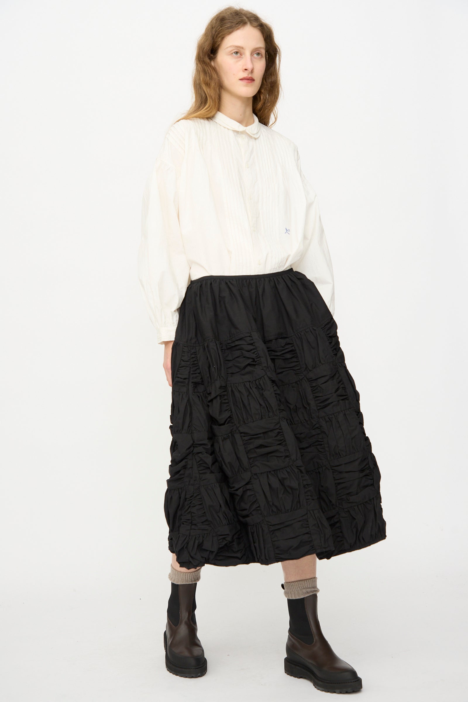 A person stands against a plain white background wearing the Story mfg. Patchwork Foxglove Skirt in Interstellar, paired with a loose white blouse, beige socks, and dark ankle boots.