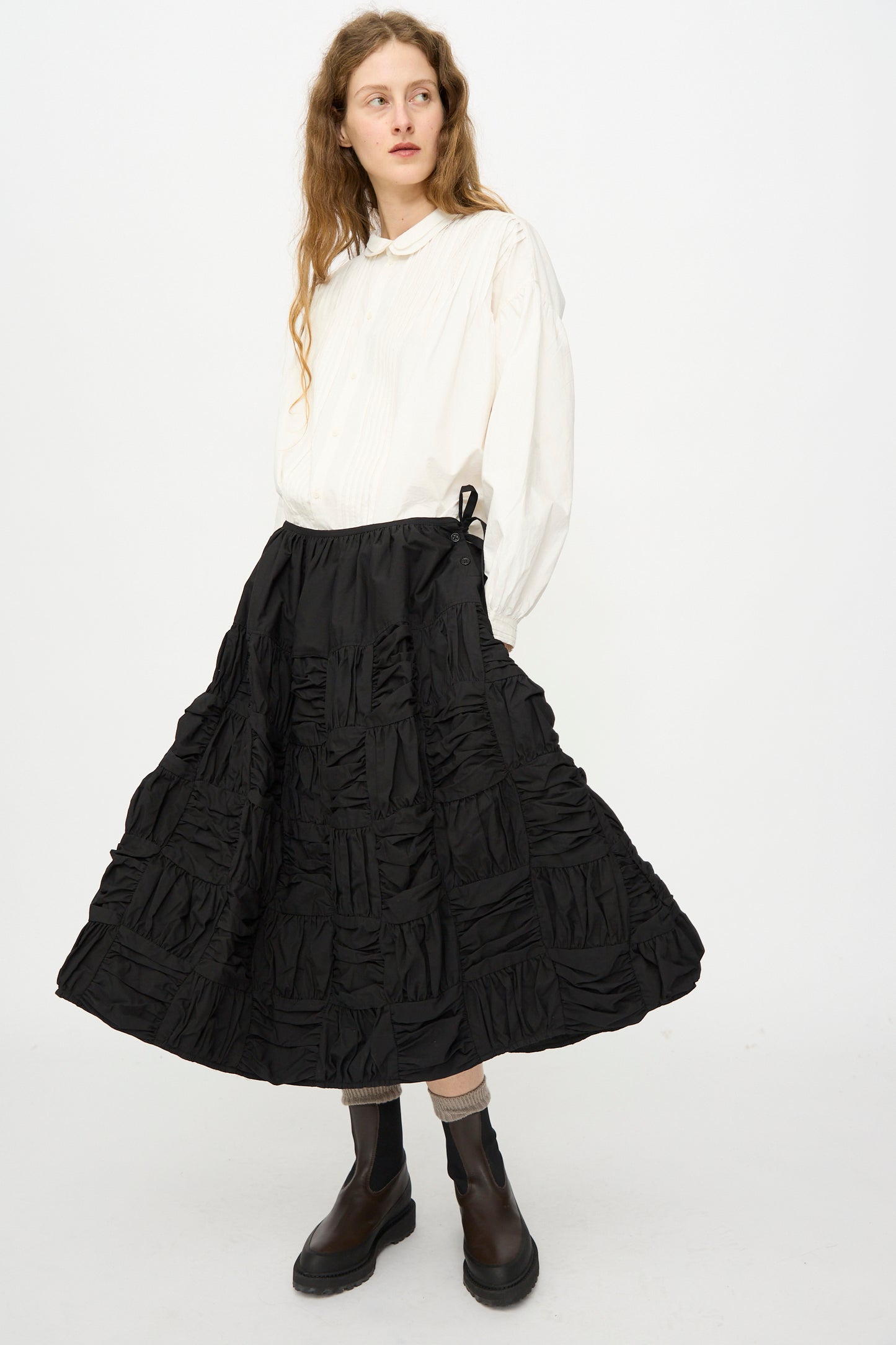 A person wears a loose white blouse, Story mfg. Patchwork Foxglove Skirt in Interstellar, black socks, and black ankle boots, standing against a plain white background.