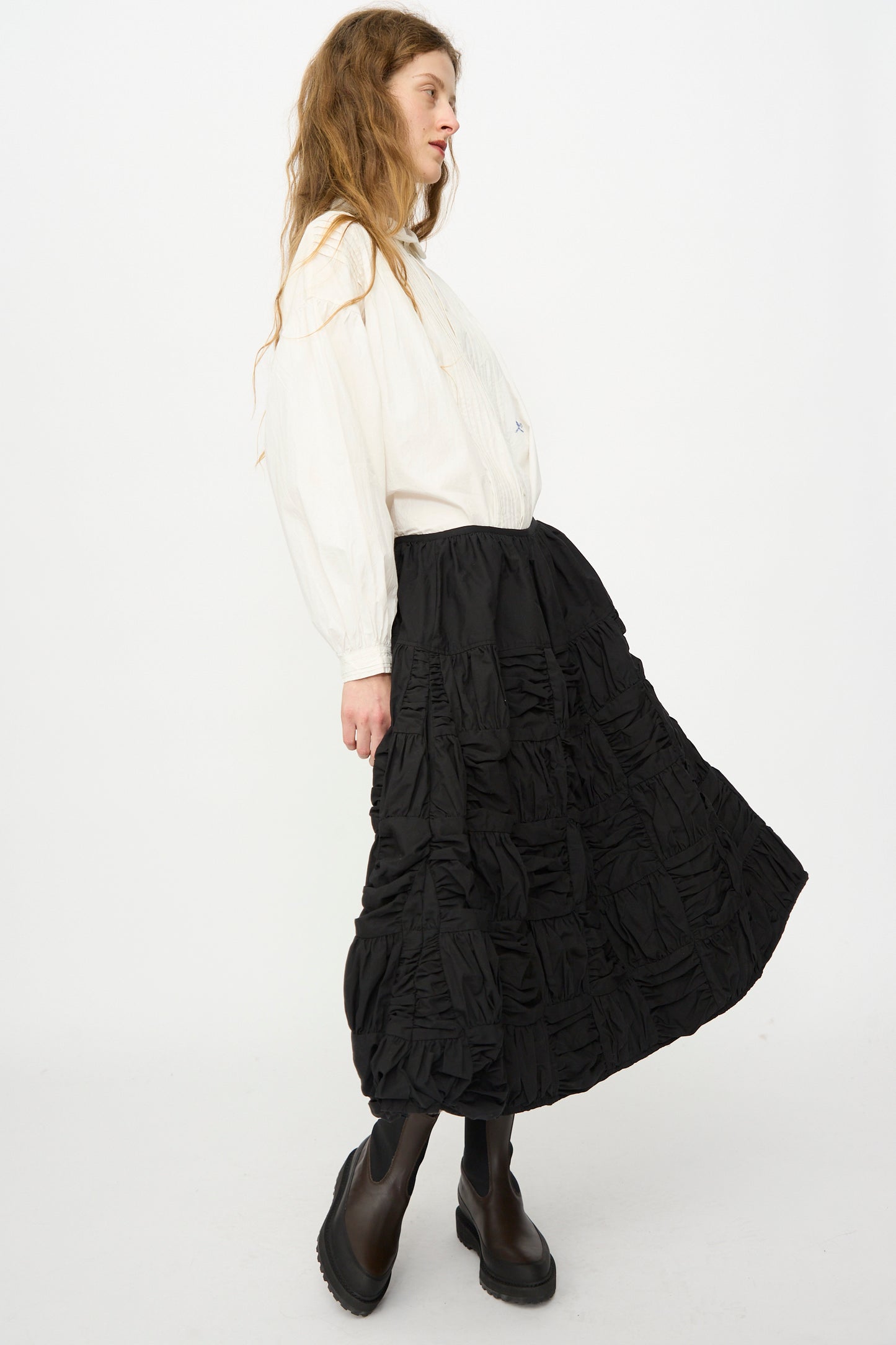 A woman stands sideways against a plain white background, wearing a white long-sleeve blouse, black boots, and the Story mfg. Patchwork Foxglove Skirt in Interstellar.