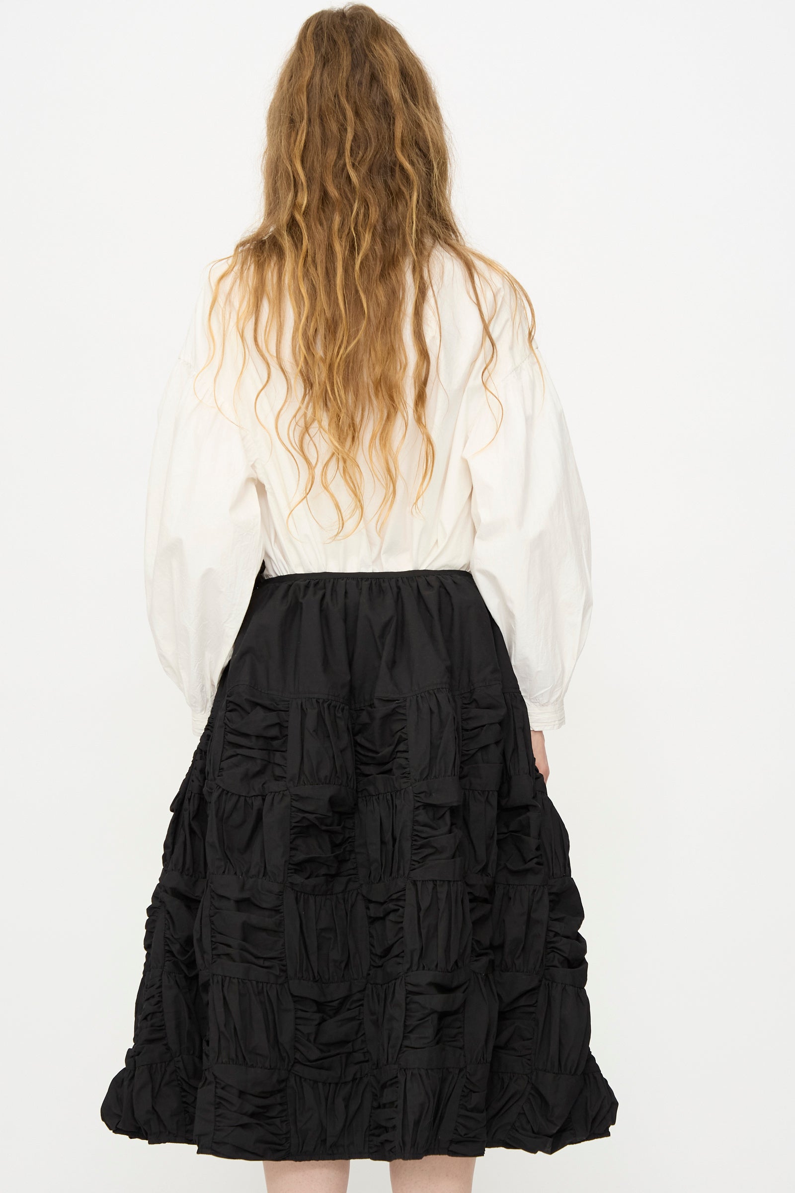 A person with long wavy hair wearing a white long-sleeve blouse and the Story mfg. Patchwork Foxglove Skirt in Interstellar stands facing away from the camera against a plain white background.