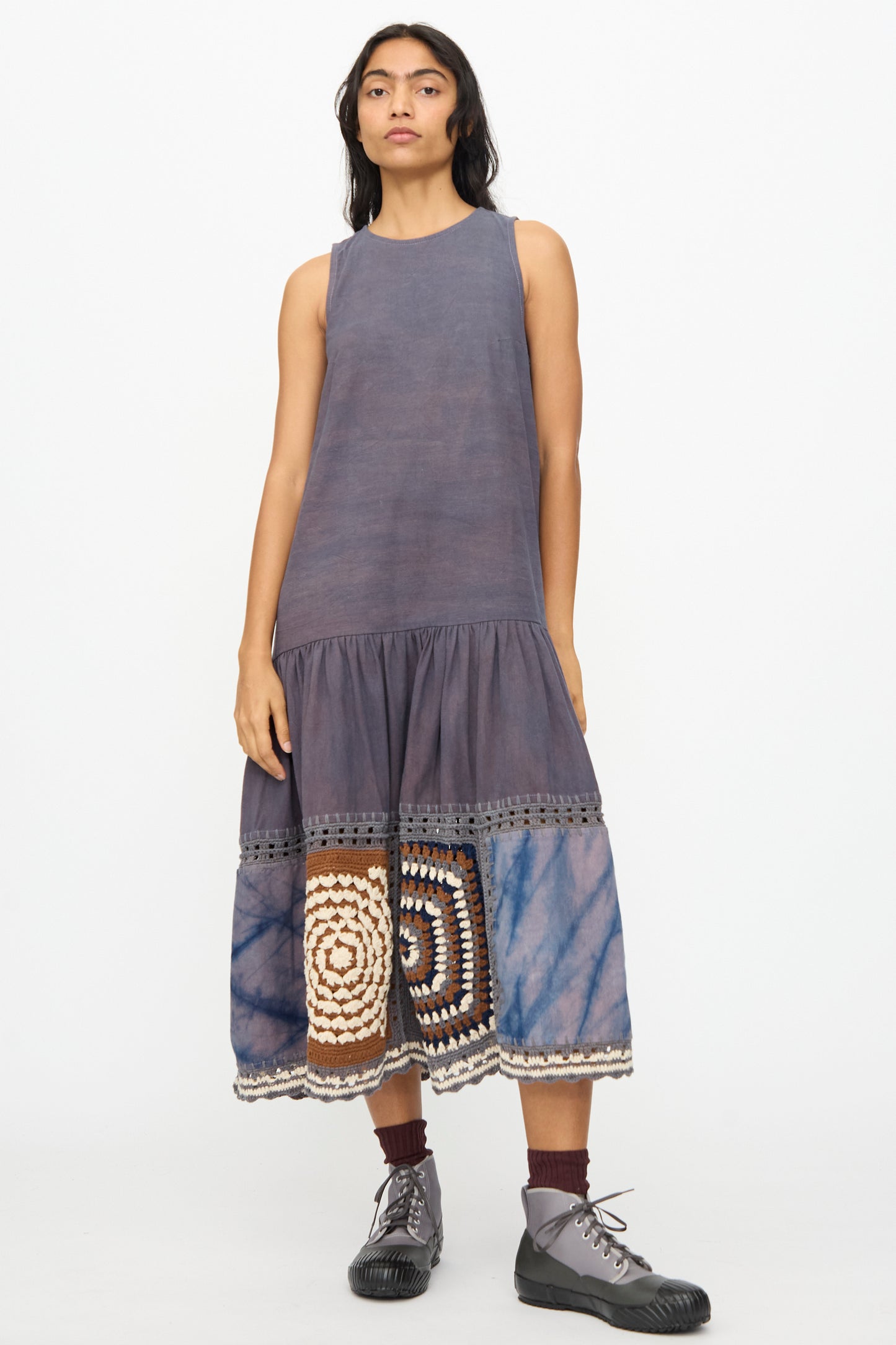 A person wears the Story mfg. Snowdrop Dress in Cosmic Crochet, a sleeveless midi with hand crochet patchwork and tie-dye near the hem, paired with brown socks and gray lace-up shoes.