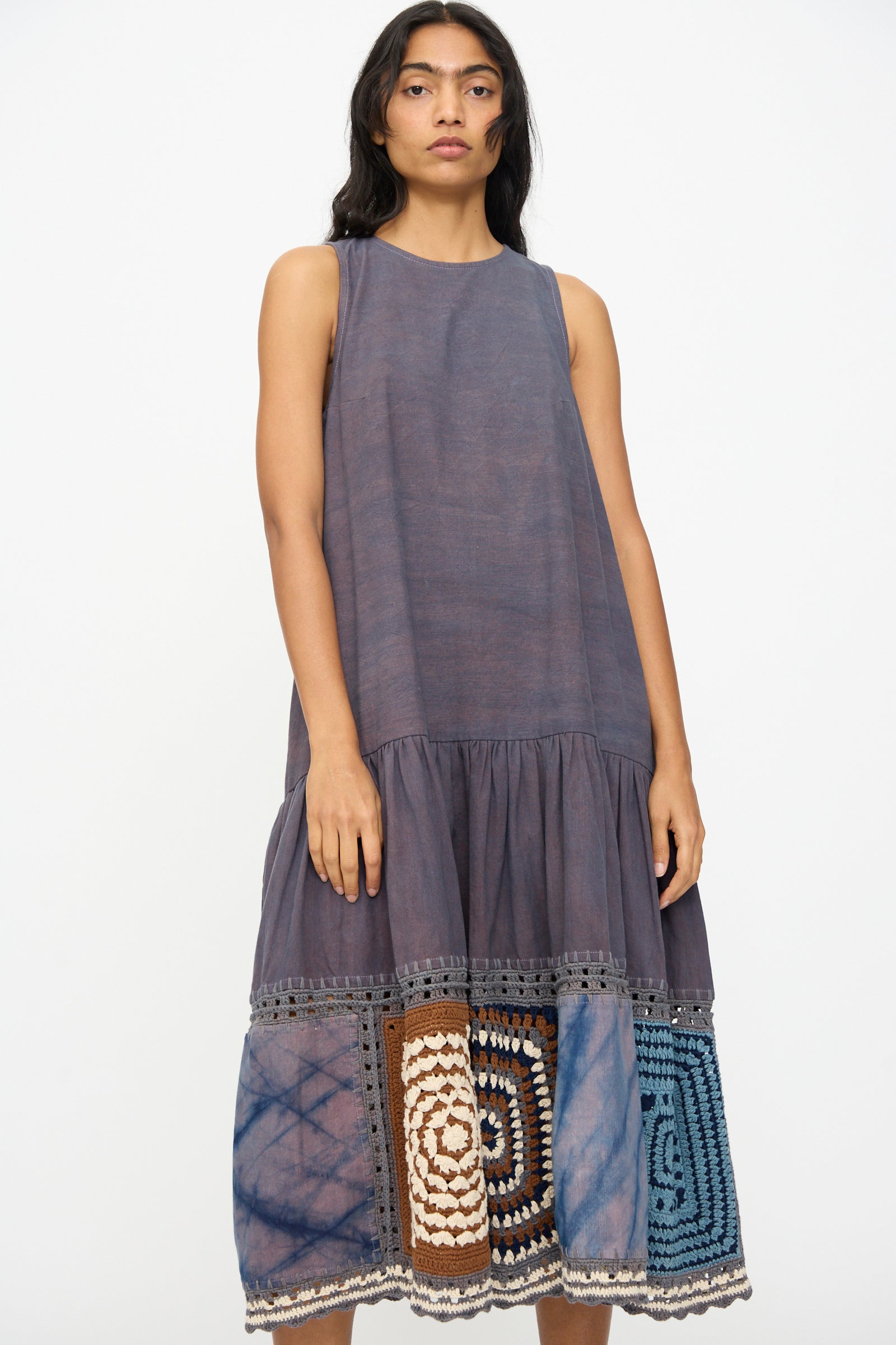 A woman wears the Story mfg. Snowdrop Dress in Cosmic Crochet, a sleeveless purple midi with a gathered skirt and hand-crochet patchwork hem in blue, brown, and cream.