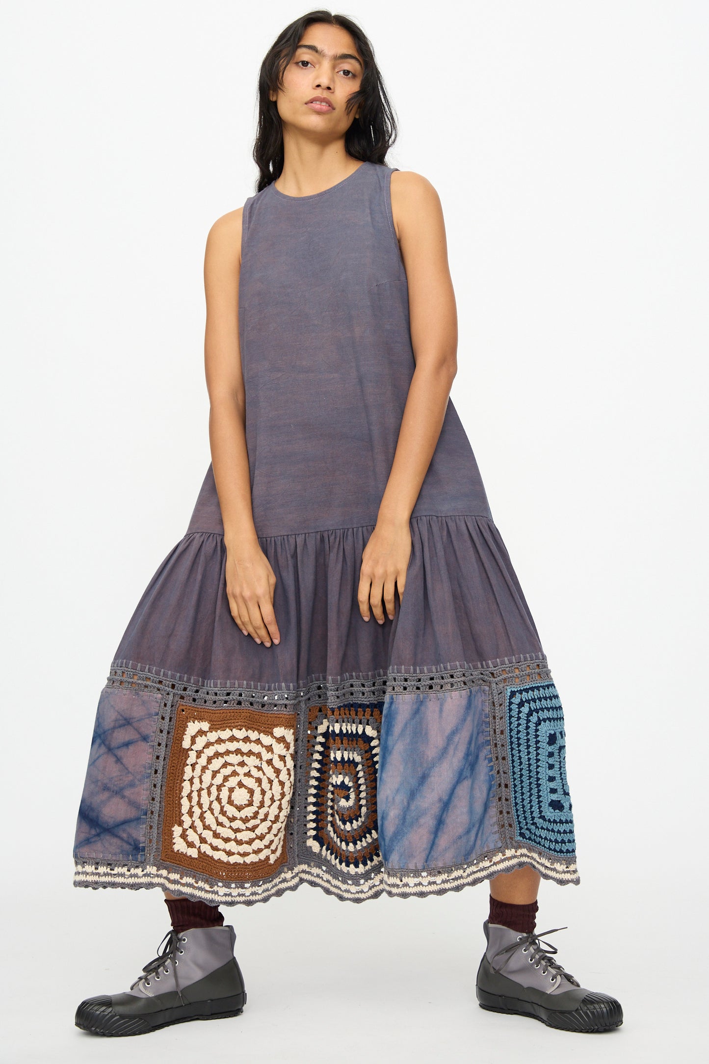 A woman wears the Story mfg. Snowdrop Dress in Cosmic Crochet—a sleeveless purple midi dress with a hand-crochet patchwork hem—paired with brown socks and gray lace-up boots, standing against a white background.