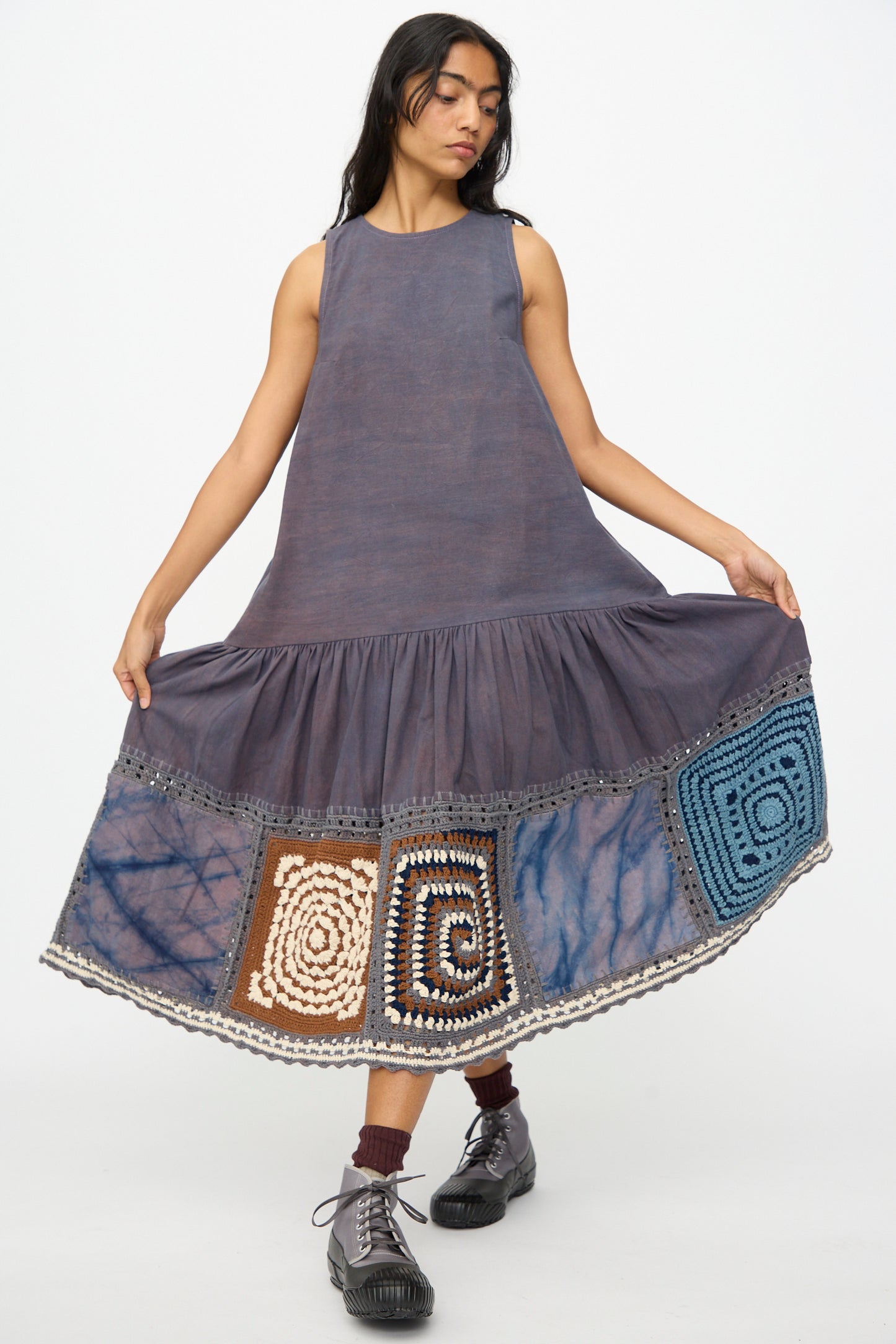 A woman models the Story mfg. Snowdrop Dress in Cosmic Crochet—a sleeveless purple midi dress with hand crochet patchwork near the hem—paired with dark socks and lace-up shoes, set against a plain background.