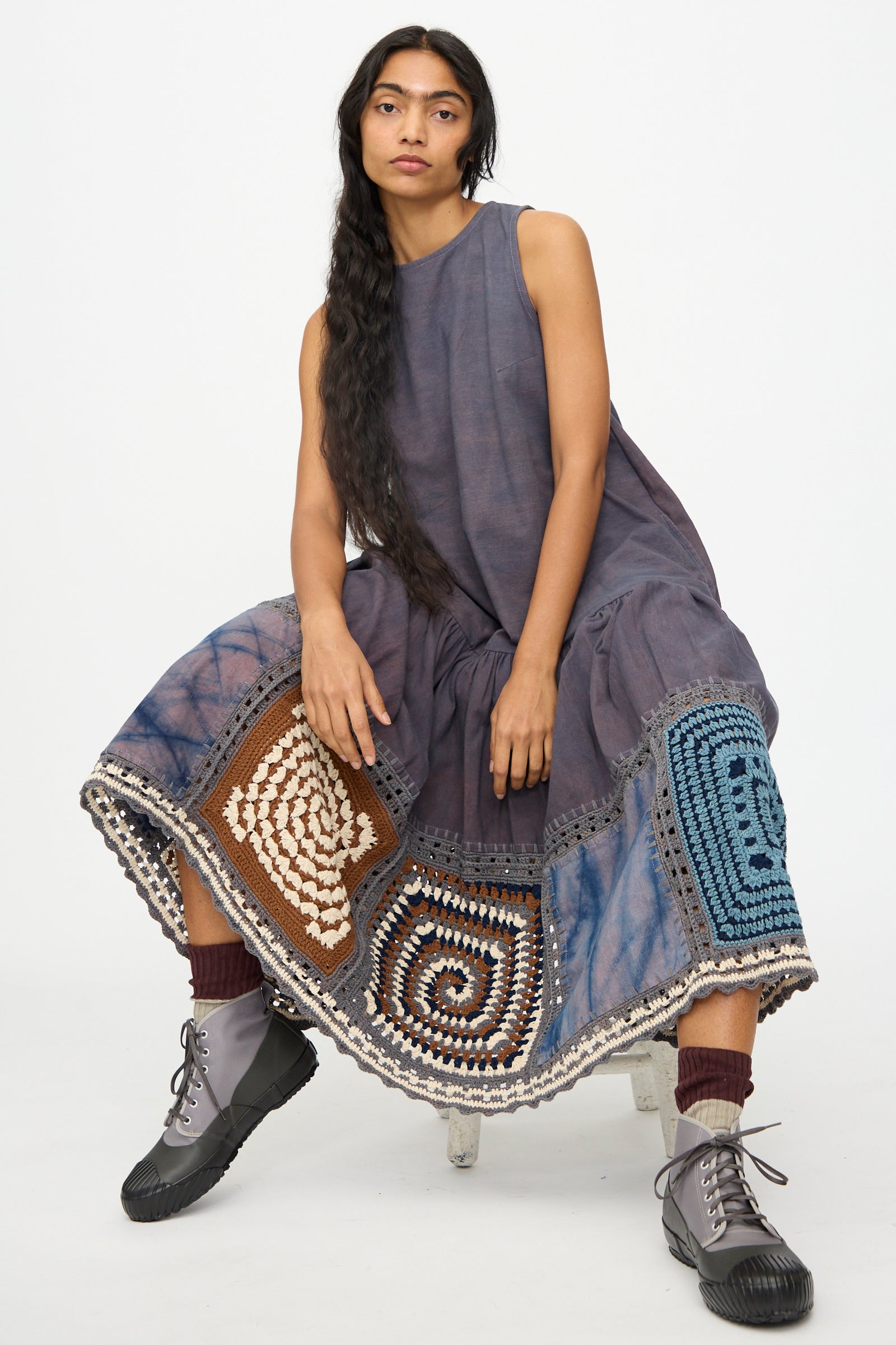 A woman sits on a stool against a plain white background wearing the Story mfg. Snowdrop Dress in Cosmic Crochet, sleeveless with hand crochet patchwork at the hem, paired with dark socks and gray lace-up boots.