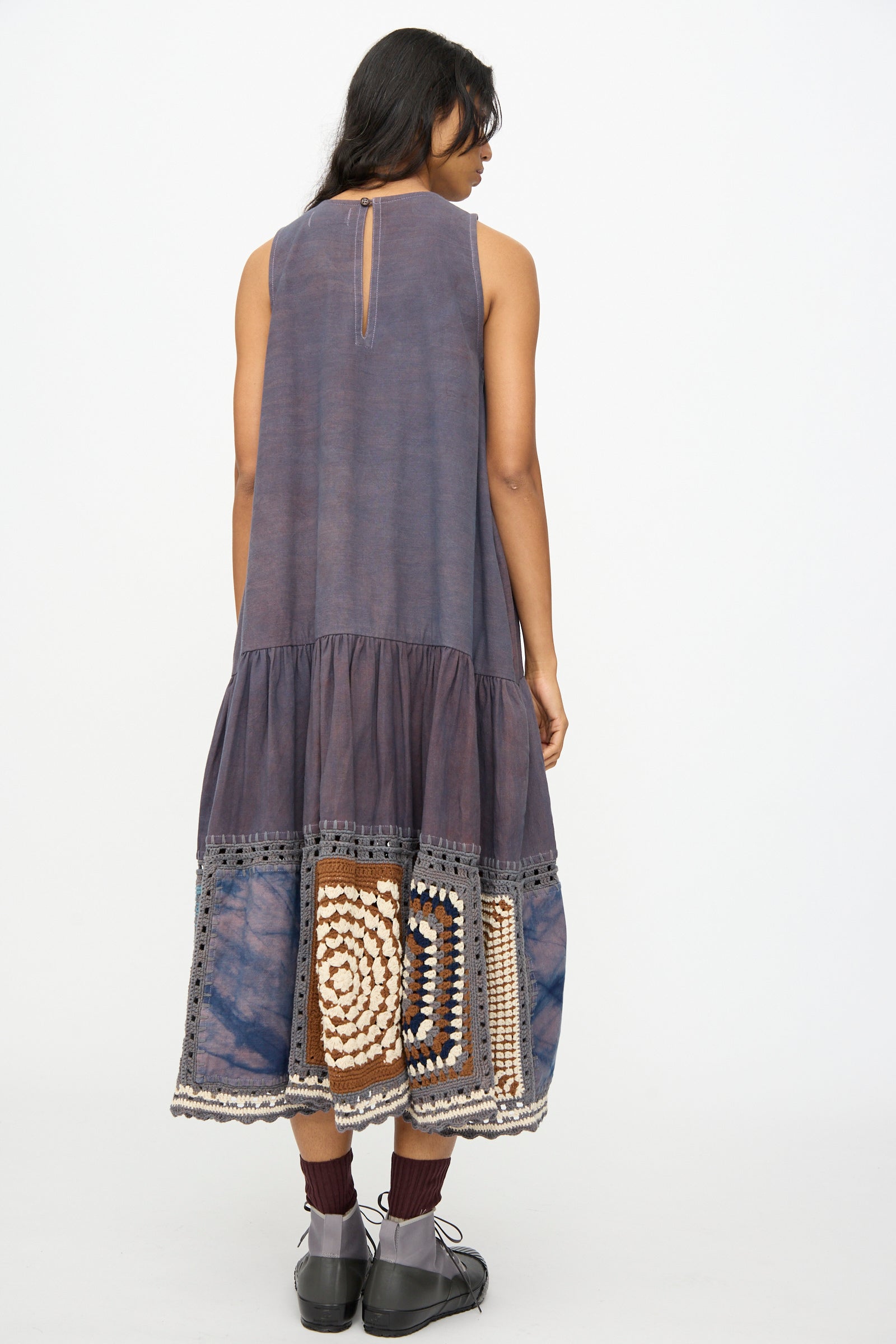 A person faces away wearing the Story mfg. Snowdrop Dress in Cosmic Crochet—a purple sleeveless midi dress with hand crochet patchwork near the hem—paired with black shoes and maroon socks.