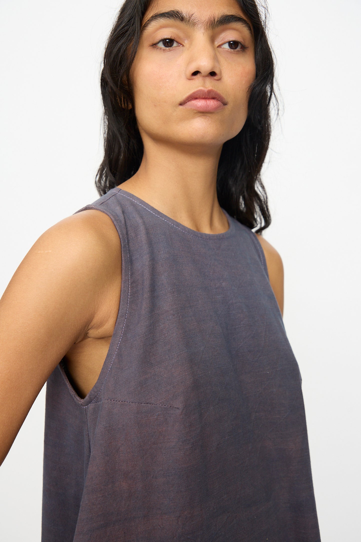 A woman with long dark hair wears the Story mfg. Snowdrop Dress in Cosmic Crochet—a sleeveless midi dress—in faded purple, standing against a plain light background and gazing slightly upward.