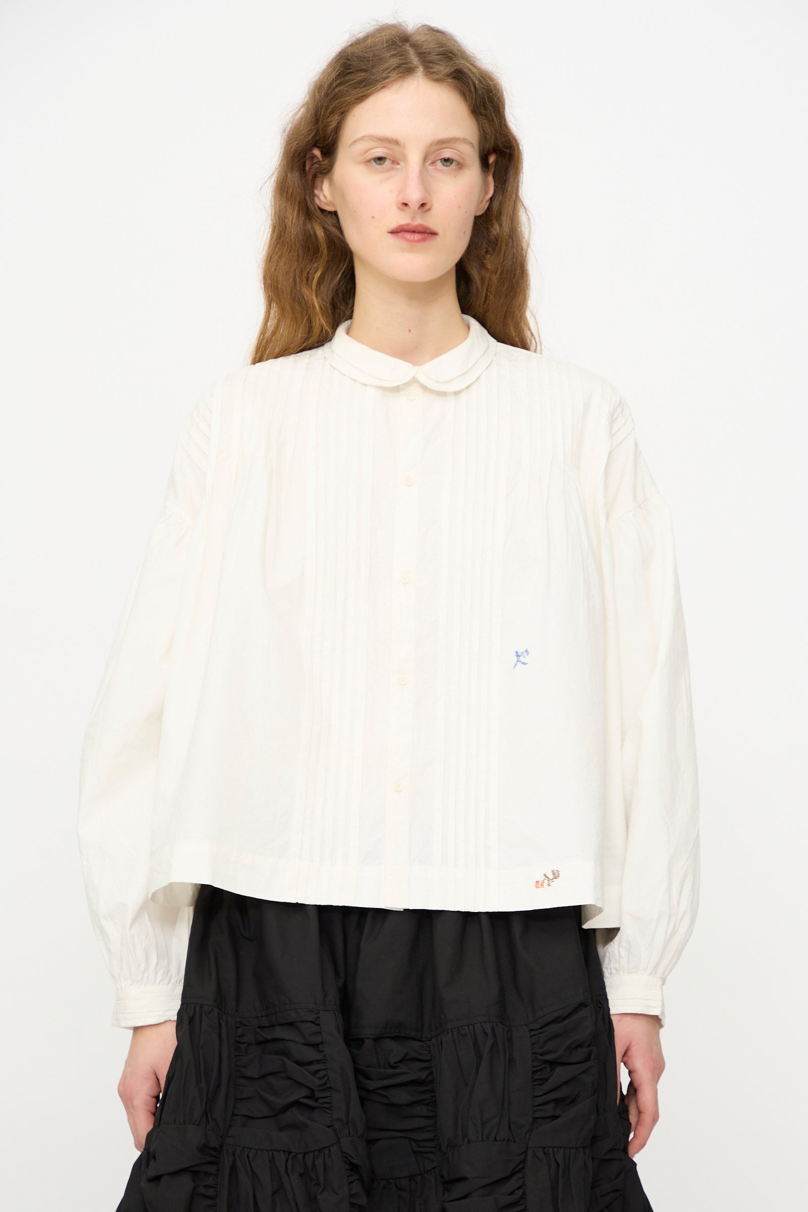 A woman with long brown hair wears the Story mfg. Still Shirt in Interrupted Embroidery, a loose-fitting organic cotton blouse with a Peter Pan collar, paired with a black skirt against a plain white background.