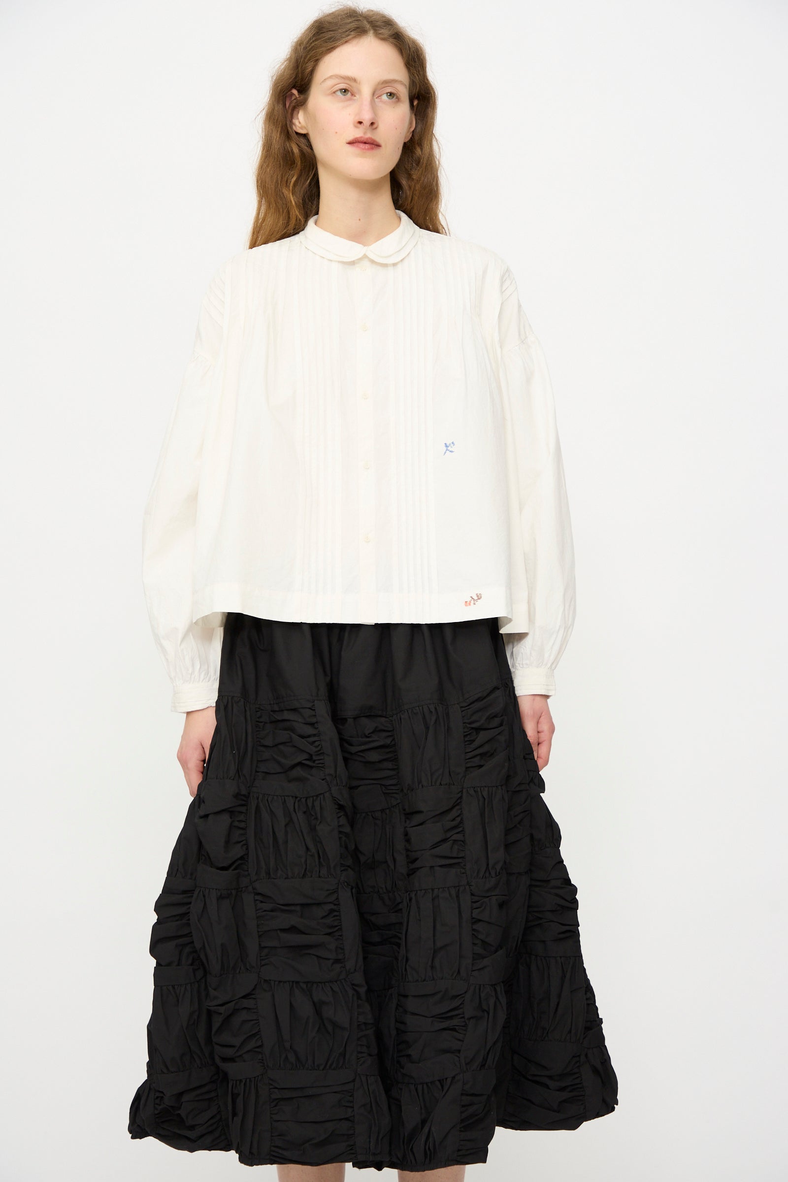 A person stands against a plain background wearing the Story mfg. Still Shirt in Interrupted Embroidery, paired with a black textured, ankle-length skirt.