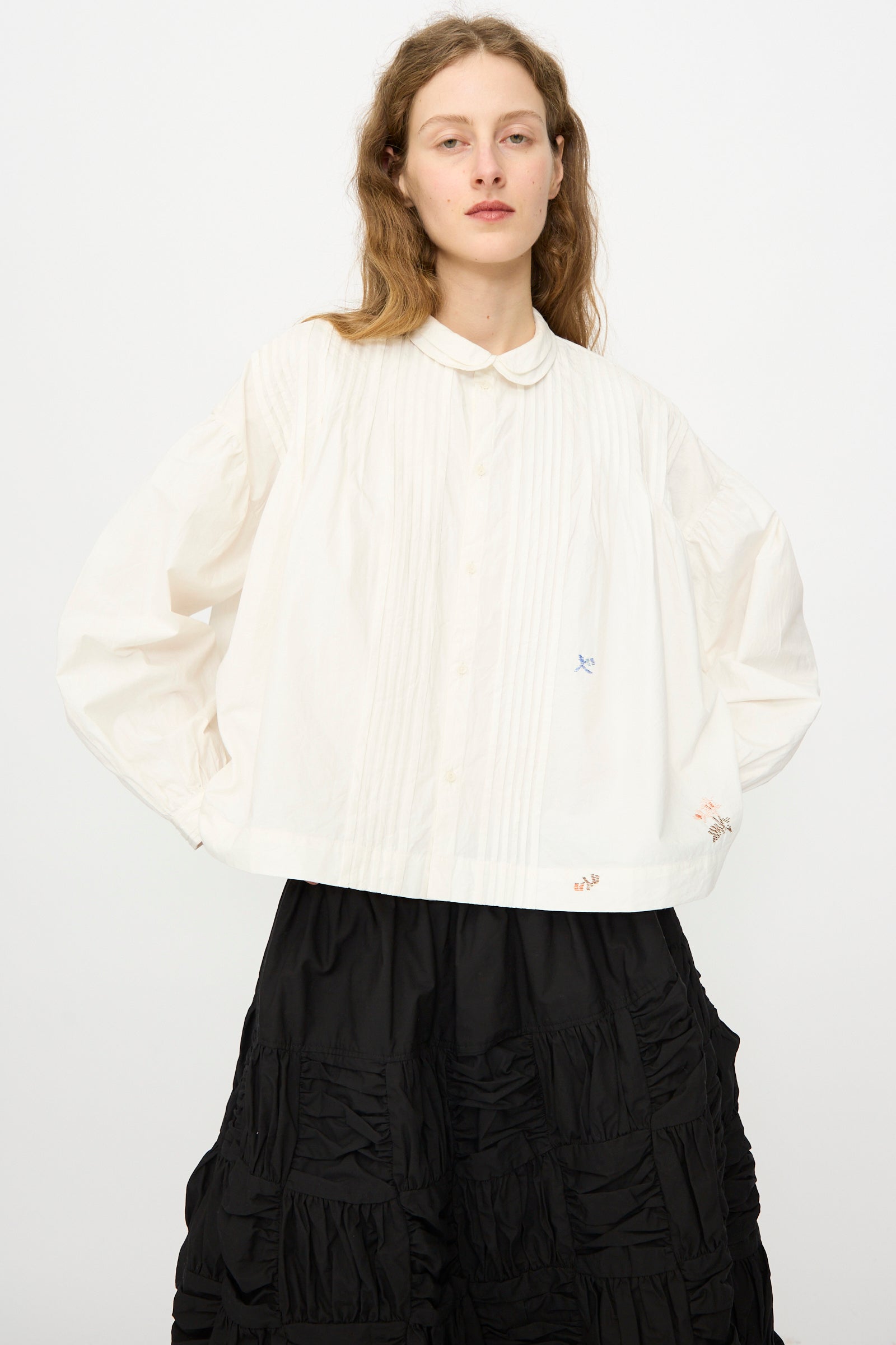 A person wears the Story mfg. Still Shirt in Interrupted Embroidery, a loose white long-sleeve blouse with subtle hand embroidery, paired with a black ruffled skirt, standing against a plain white background.