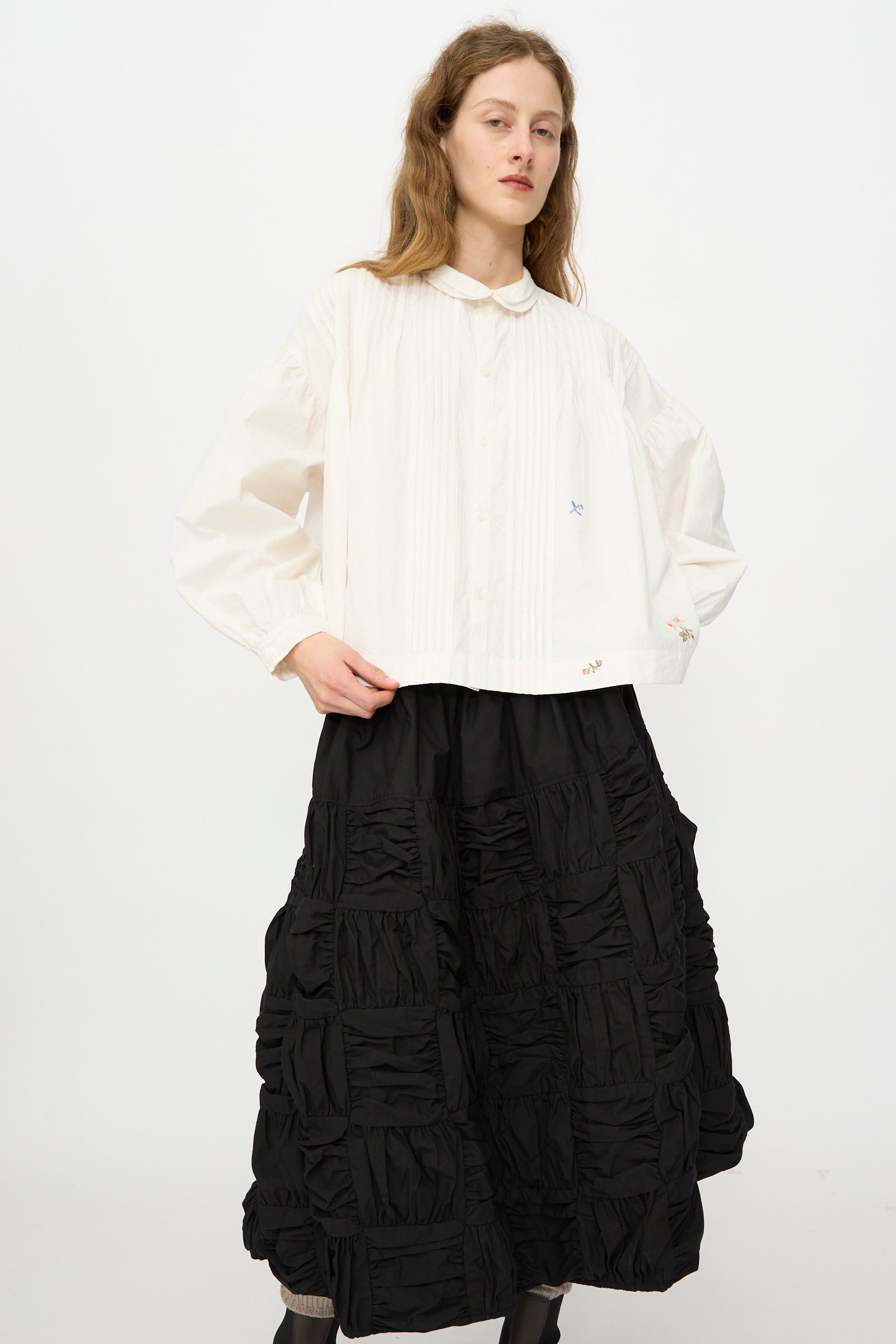A person wears the Story mfg. Still Shirt in Interrupted Embroidery, a white long-sleeve organic cotton blouse with hand embroidery, paired with a black textured voluminous skirt, standing against a plain white background.