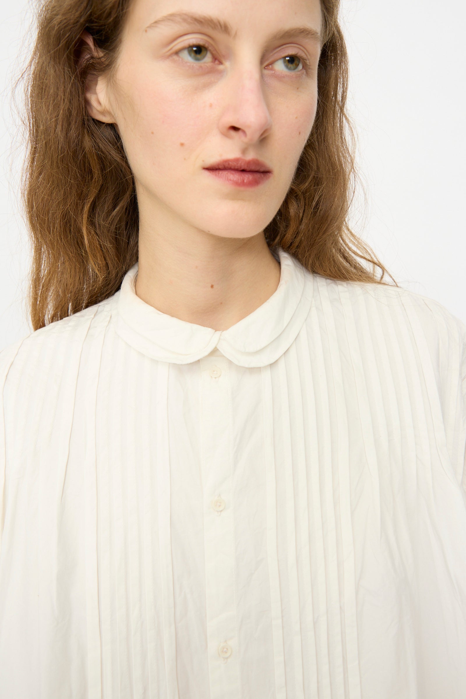A woman with light skin and long brown hair models the Story mfg. Still Shirt in Interrupted Embroidery, a white pleated organic cotton blouse with a rounded collar, looking slightly to the side against a plain background.