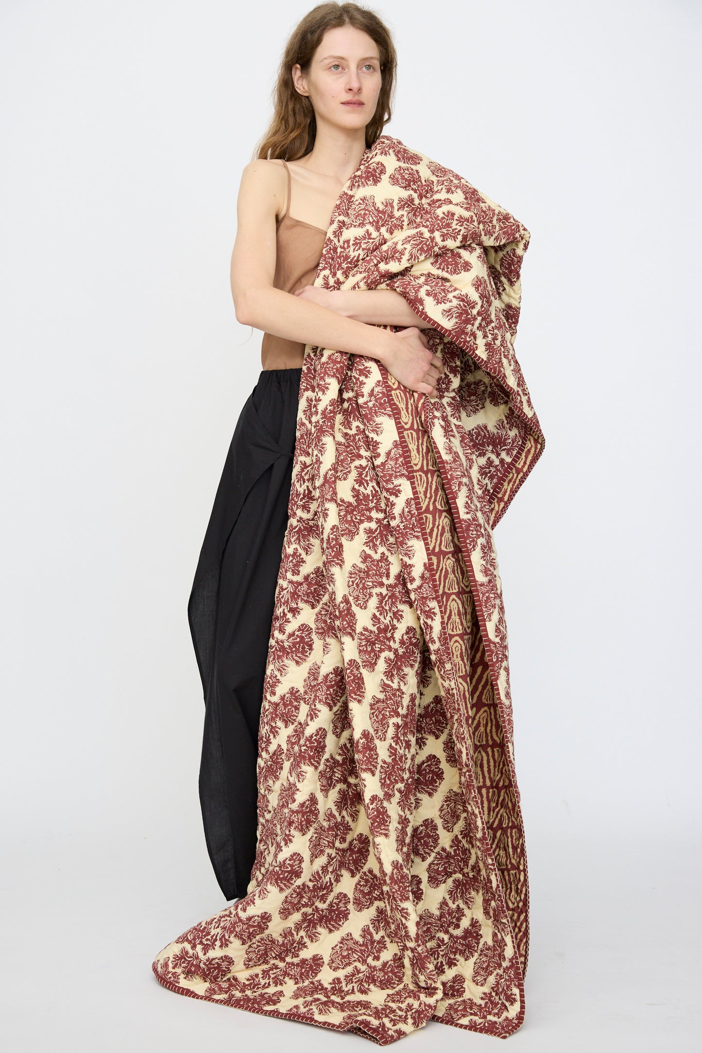A person stands against a white background, draped in the Studio Ford Wild Shroom Quilt 100 x 108", a large cream and maroon block-printed percale cotton blanket, wearing a tan top and black skirt.