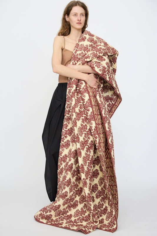 A person stands against a white background, draped in the Studio Ford Wild Shroom Quilt 100 x 108", a large cream and maroon block-printed percale cotton blanket, wearing a tan top and black skirt.