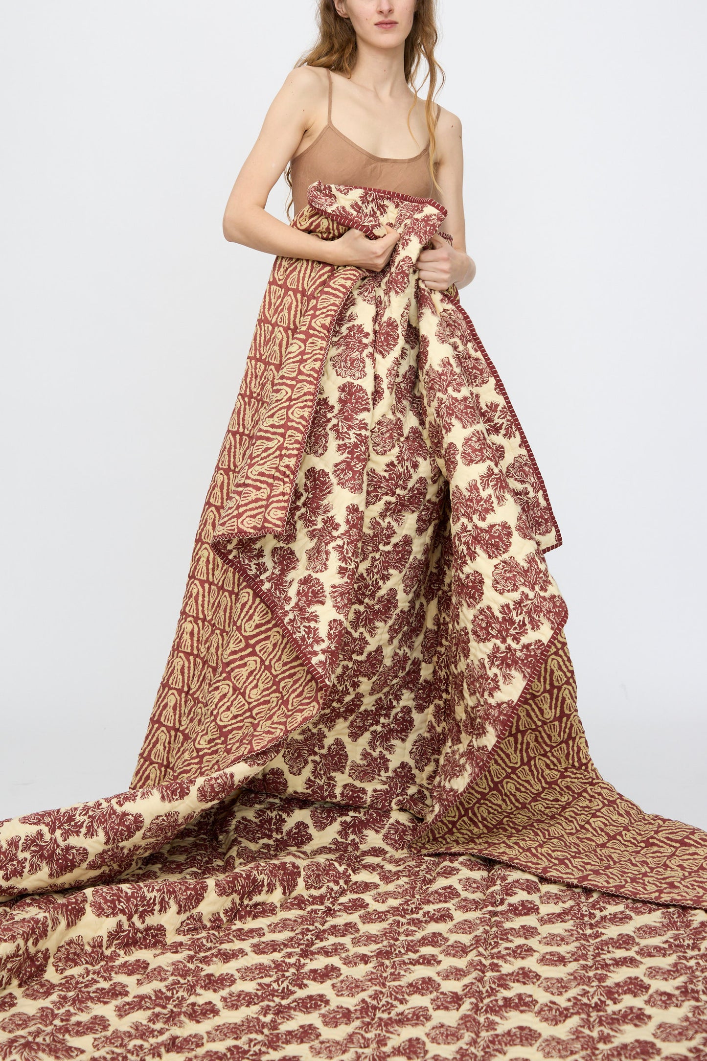 A woman in a tan camisole displays the Studio Ford Wild Shroom Quilt (100 x 108"), featuring flowing, block-printed red floral and abstract patterns on percale cotton, set against a plain white background.