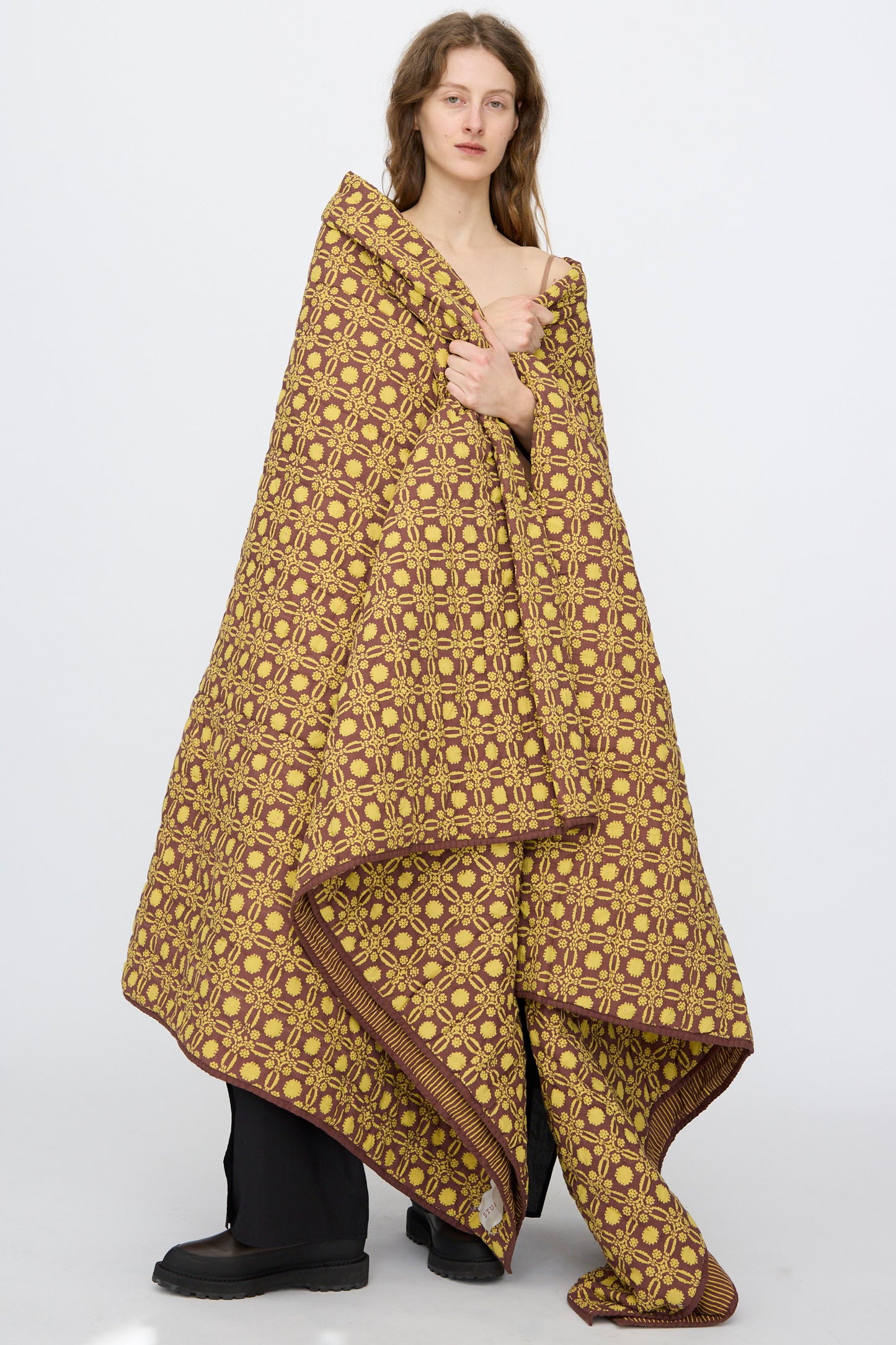 A person stands wrapped in the Studio Ford Thatcher Quilt 100 x 108", featuring a yellow and brown geometric design, wearing black pants and shoes against a plain white background.