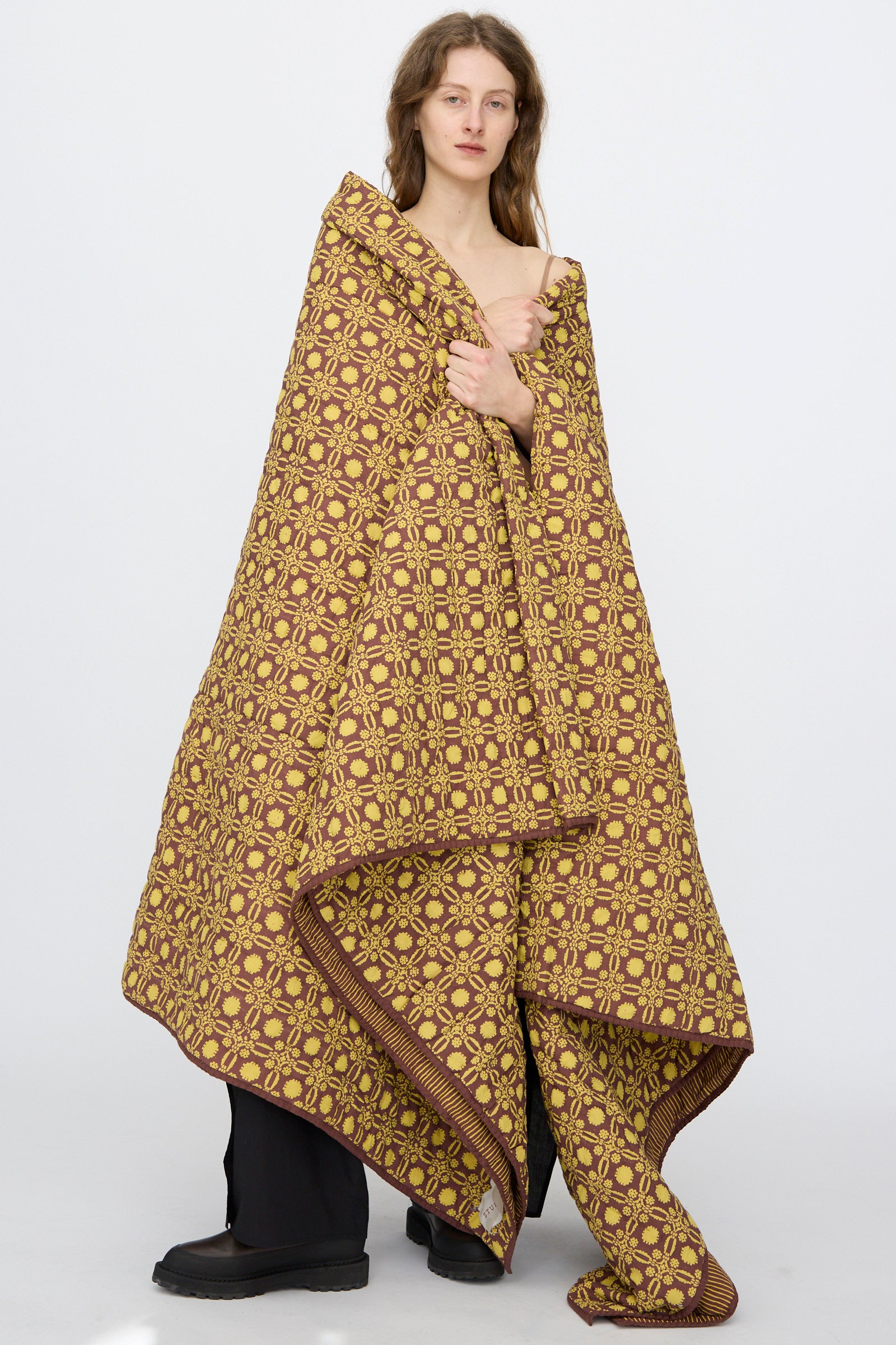 A person stands wrapped in the Studio Ford Thatcher Quilt 100 x 108", featuring a yellow and brown geometric design, wearing black pants and shoes against a plain white background.