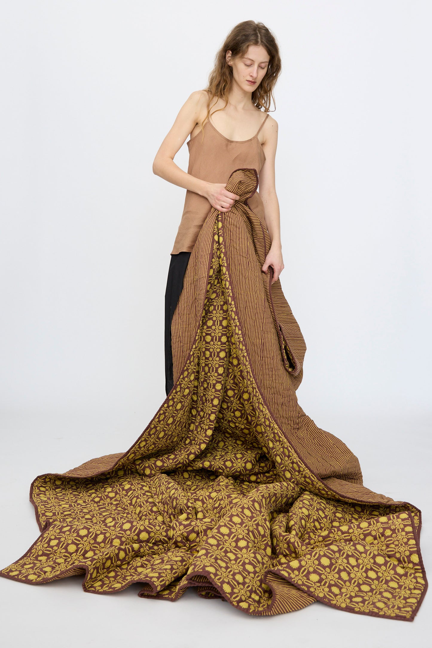 A woman in a brown tank top holds up the Studio Ford Thatcher Quilt (100 x 108"), featuring handmade block-printed percale cotton in brown and yellow patterns, with the fabric draping onto the floor.