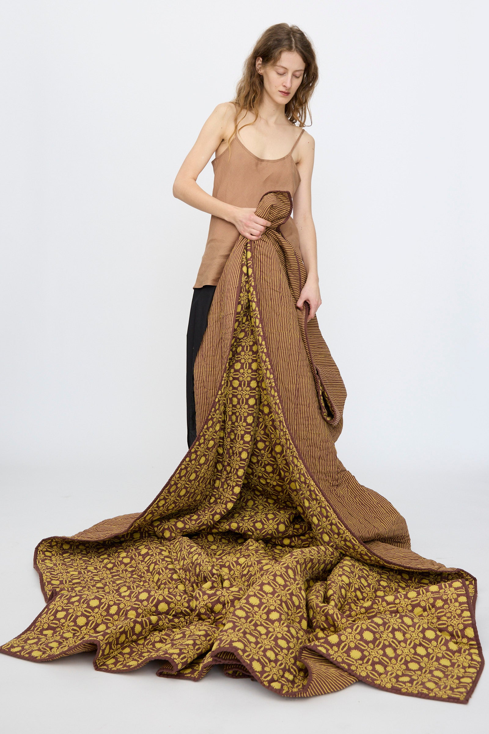 A woman in a brown tank top holds up the Studio Ford Thatcher Quilt (100 x 108"), featuring handmade block-printed percale cotton in brown and yellow patterns, with the fabric draping onto the floor.