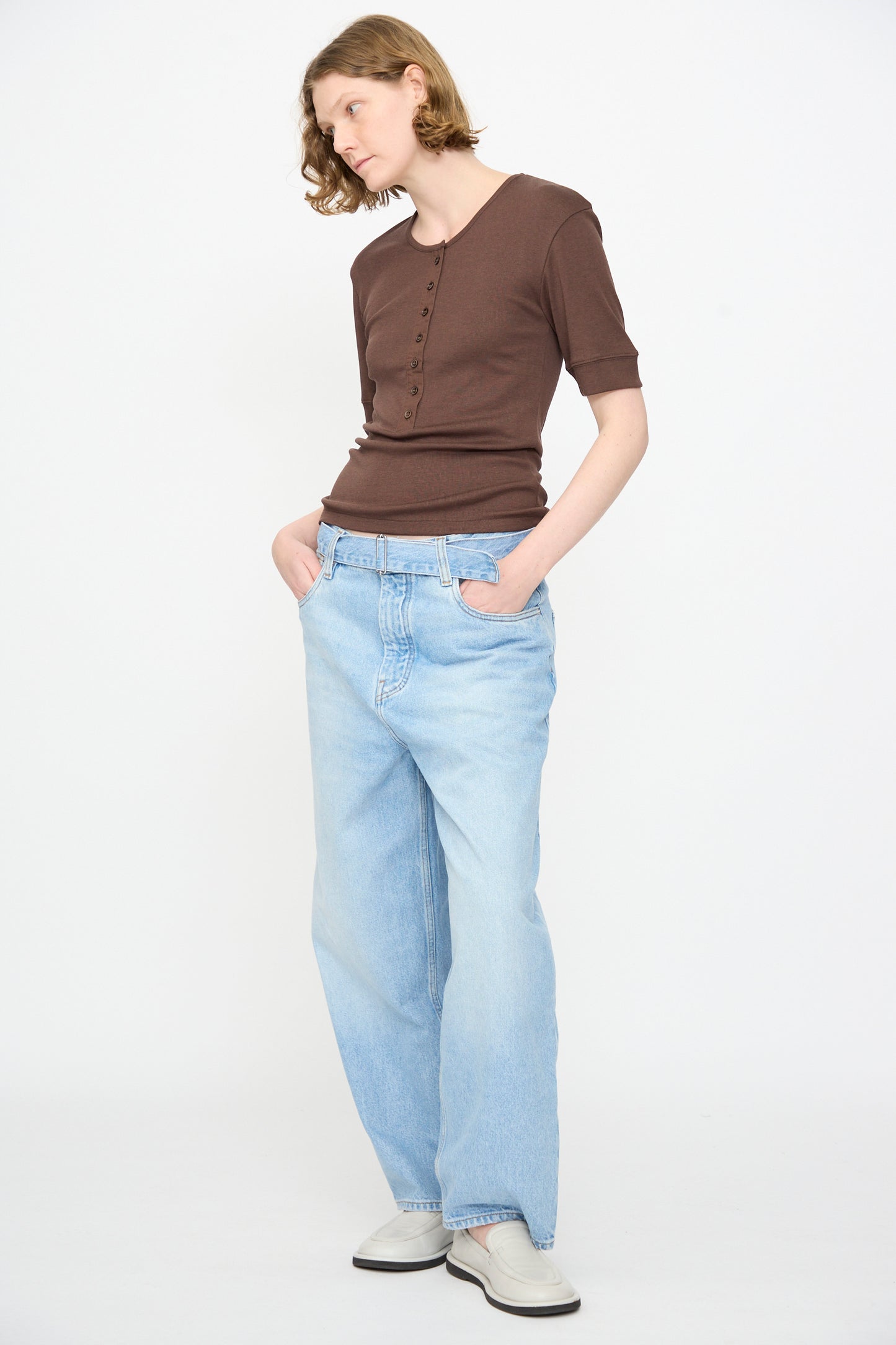 Person wearing the Studio Nicholson Sorin Short Sleeve Henley in Espresso, light blue loose jeans, and white shoes stands with hands in pockets against a plain white background.