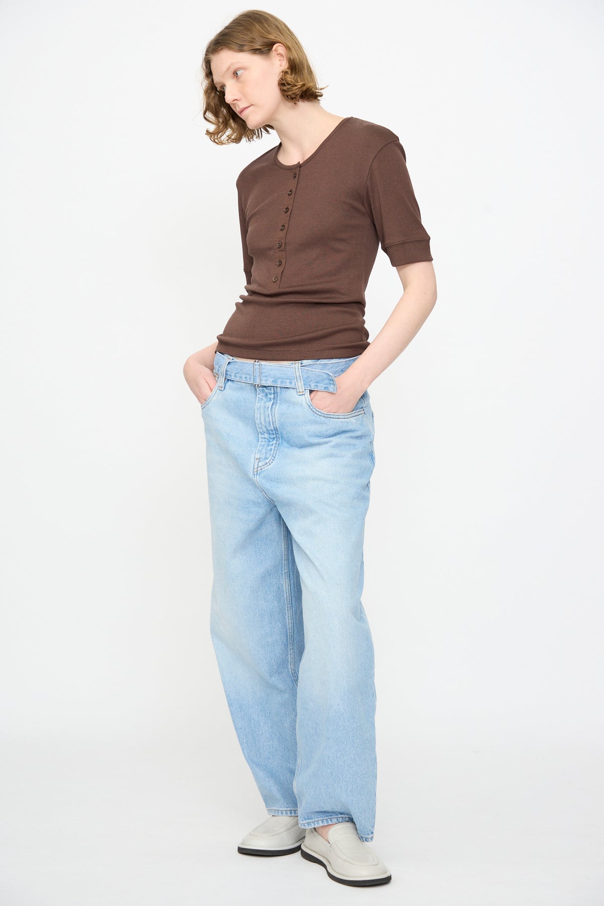 Person wearing the Studio Nicholson Sorin Short Sleeve Henley in Espresso, light blue loose jeans, and white shoes stands with hands in pockets against a plain white background.