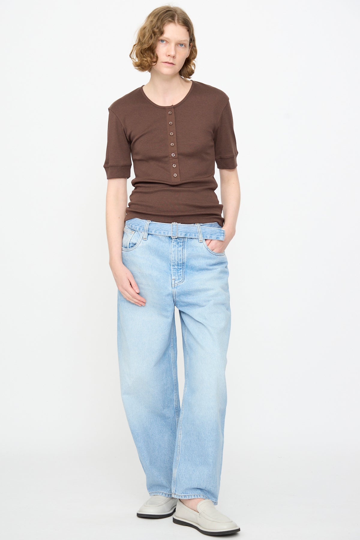 A person with short wavy hair wearing the Studio Nicholson Sorin Short Sleeve Henley in Espresso, light blue loose-fit jeans, and white shoes stands against a plain white background.