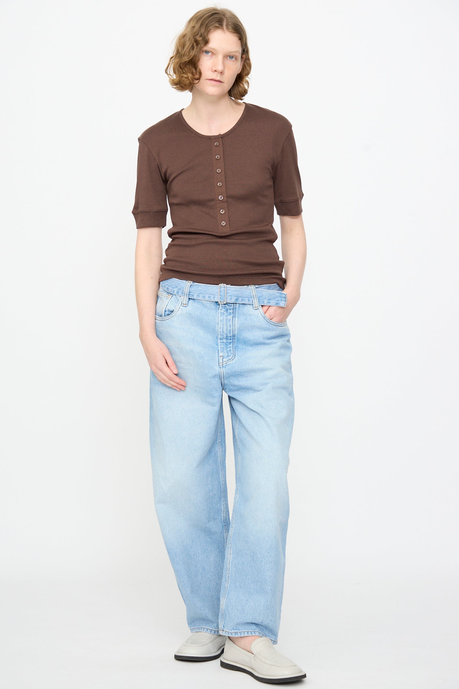 A person with short wavy hair wearing the Studio Nicholson Sorin Short Sleeve Henley in Espresso, light blue loose-fit jeans, and white shoes stands against a plain white background.