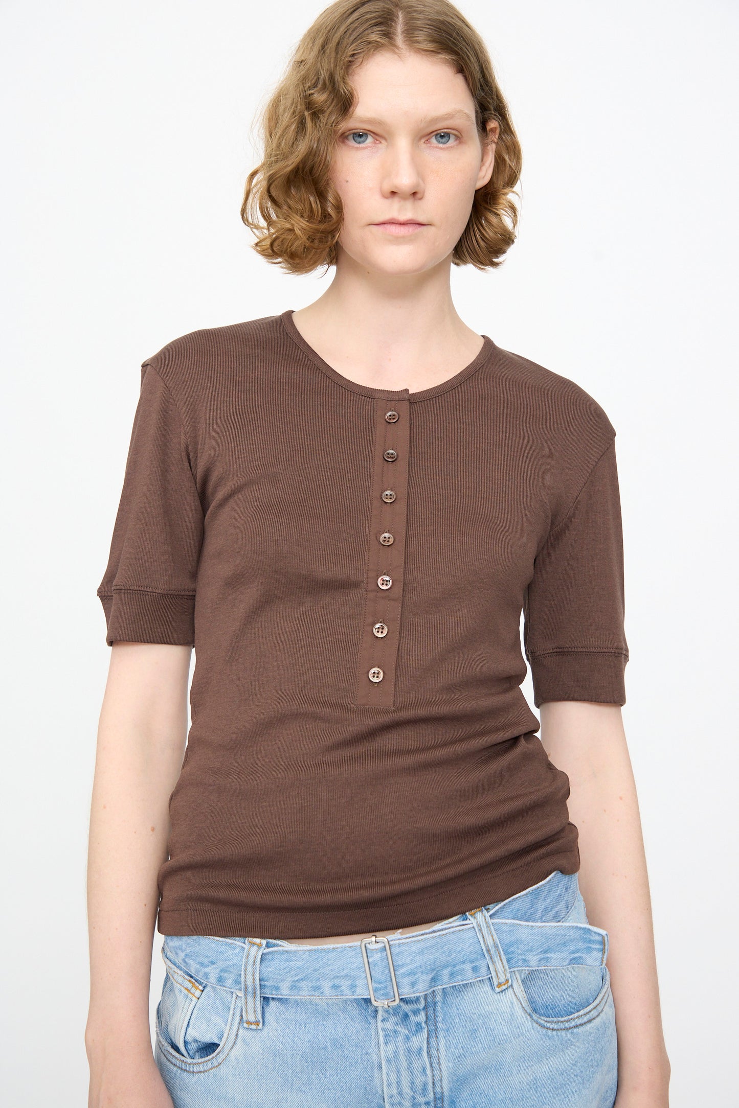 A person wearing the Studio Nicholson Sorin Short Sleeve Henley in Espresso—featuring a slim fit, ribbed cotton blend fabric, and button detailing—paired with light blue jeans, stands against a plain white background.