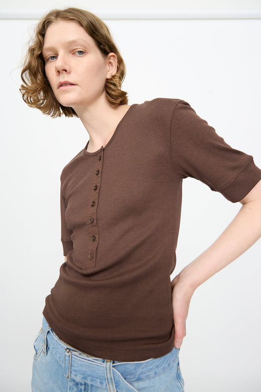 A person with short, wavy hair is wearing the Studio Nicholson Sorin Short Sleeve Henley in Espresso and light blue jeans, standing against a plain white background.