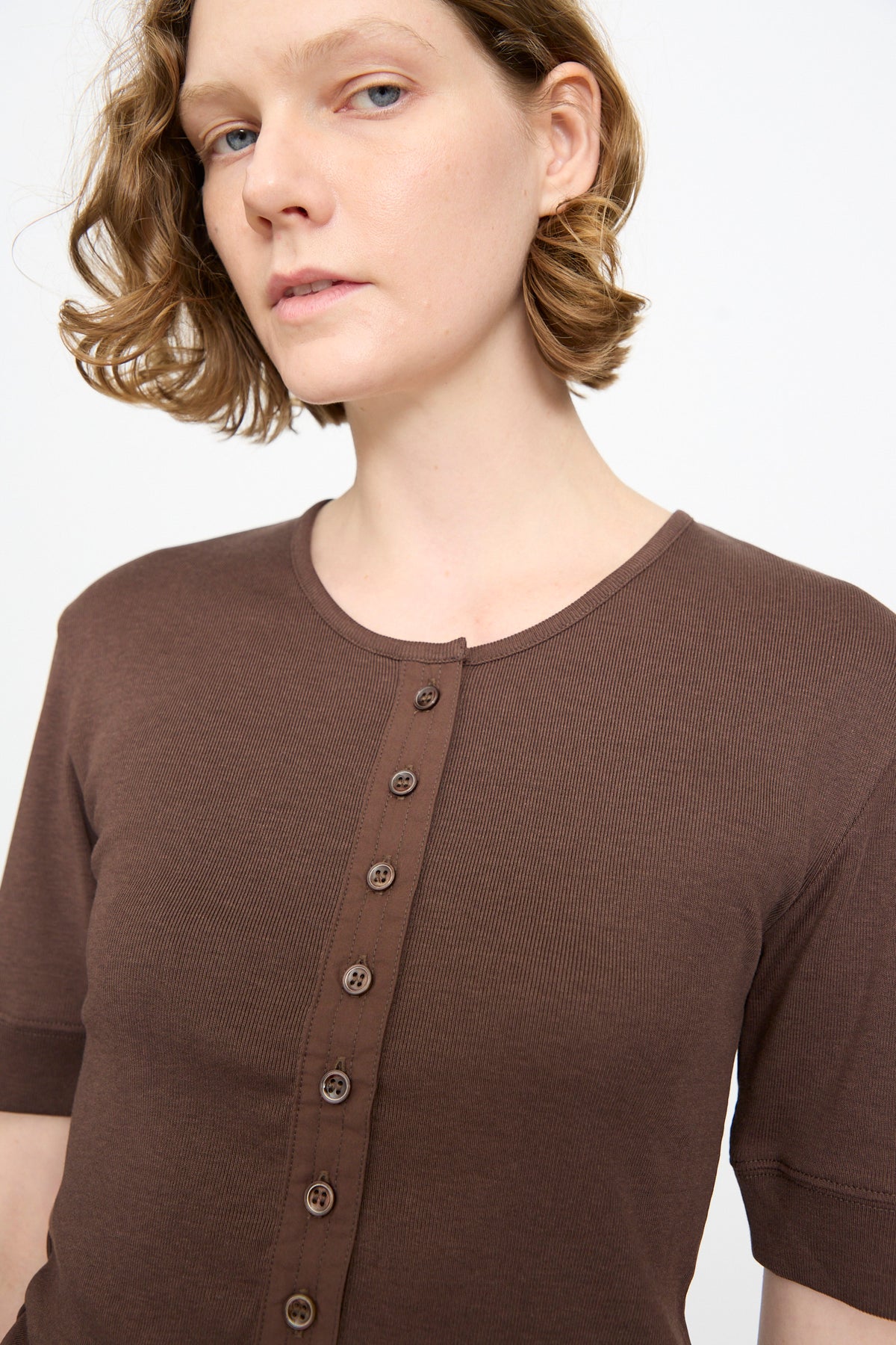 A person with short, wavy light brown hair wears the Studio Nicholson Sorin Short Sleeve Henley in Espresso and looks slightly toward the camera against a plain white background.