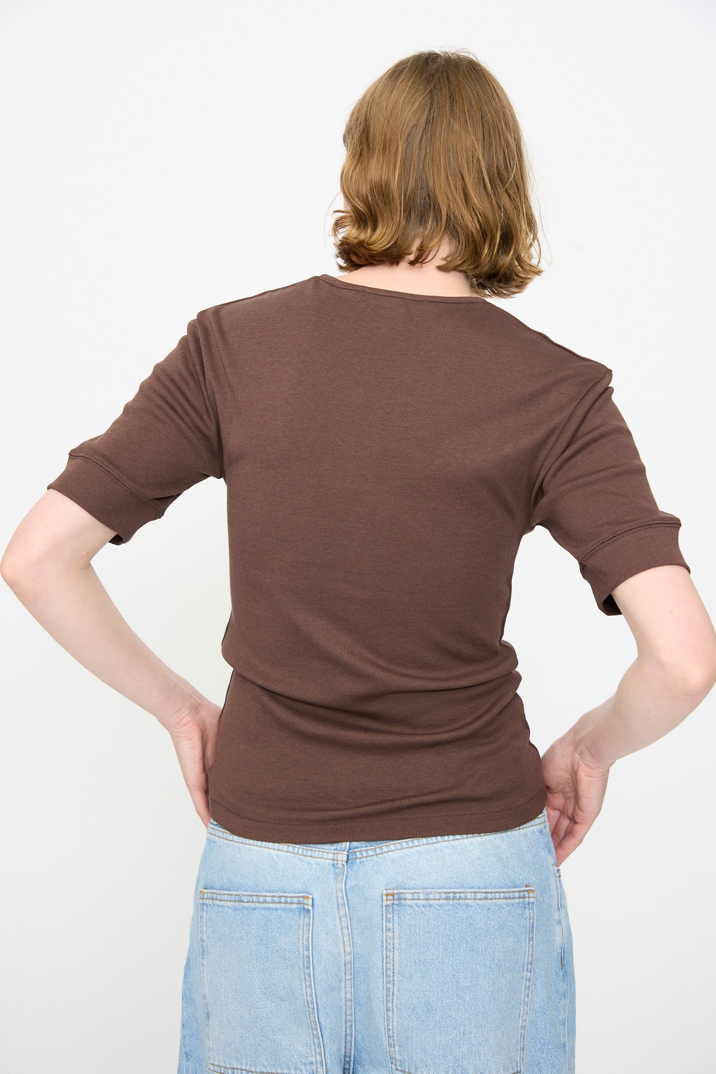 Person with short light brown hair, wearing the Studio Nicholson Sorin Short Sleeve Henley in Espresso and light blue jeans, standing with their back to the camera against a plain white background.