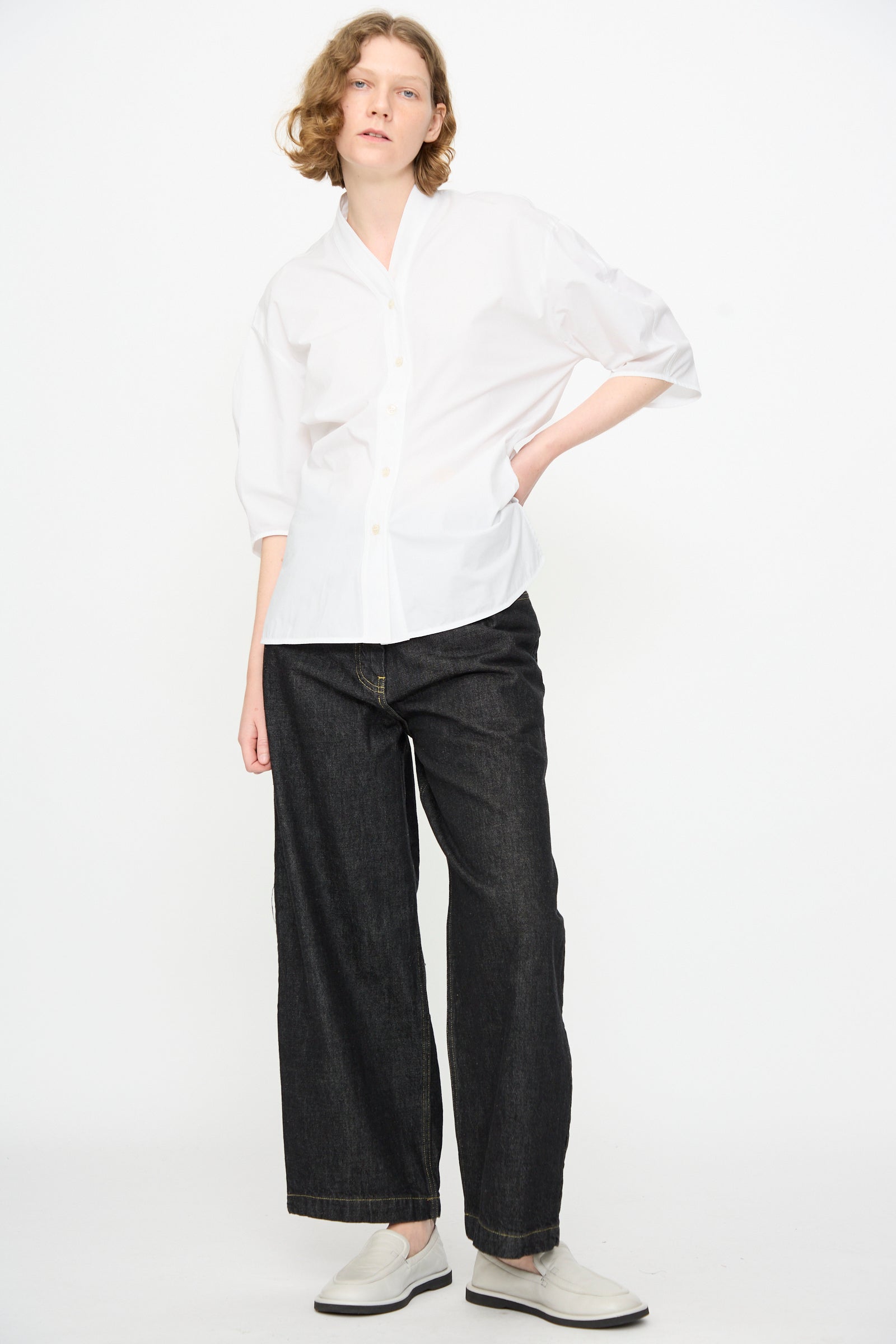 A person stands against a plain white background, wearing a white button-up shirt, the Studio Nicholson 13 oz. Denim Nieve Pant in Black—wide-leg jeans crafted from premium Italian denim—and white slip-on shoes.