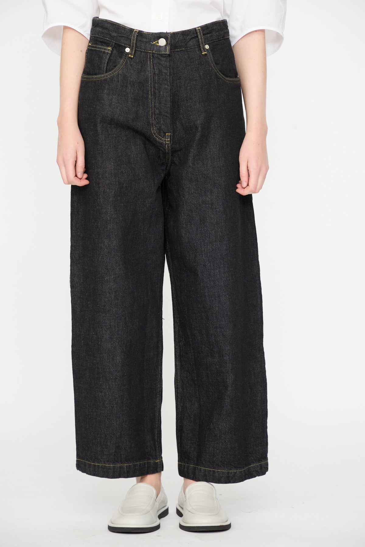 A person wearing a white shirt, Studio Nicholson 13 oz. Denim Nieve Pant in Black—relaxed, wide-leg Italian denim jeans made from organic cotton—and white slip-on shoes, standing against a plain white background.