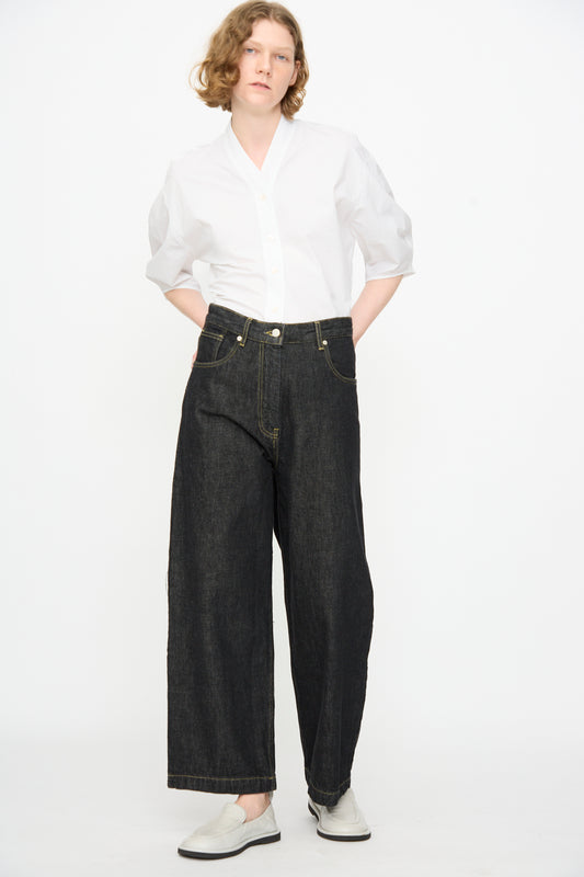 A person stands against a plain background wearing a white button-up shirt, Studio Nicholson 13 oz. Denim Nieve Pant in Black, and white shoes.