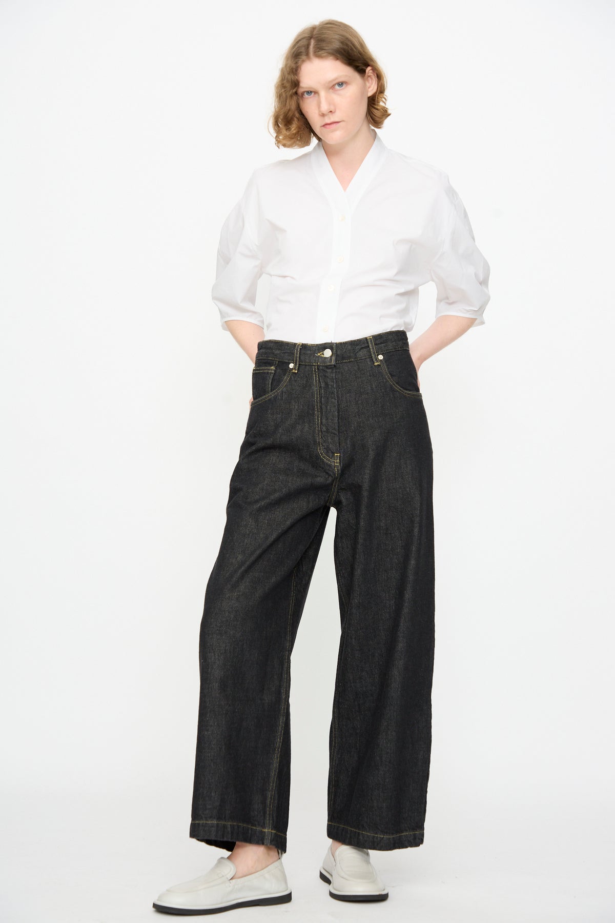 A person stands against a white background, wearing a white button-up shirt, Studio Nicholson 13 oz. Denim Nieve Pant in Black—wide-leg, relaxed Italian denim jeans made from organic cotton—and white slip-on shoes, with hands in pockets and looking at the camera.