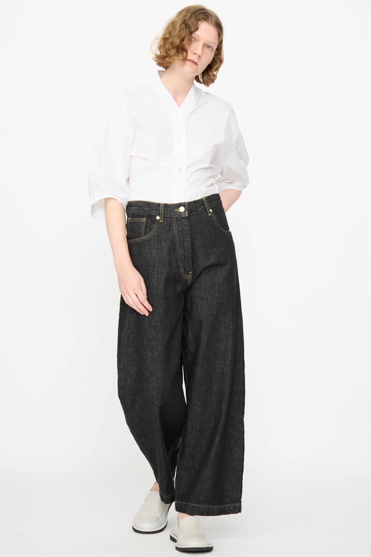 A person wearing a white button-up shirt, the Studio Nicholson 13 oz. Denim Nieve Pant in Black crafted from organic cotton, and white shoes stands against a plain white background.