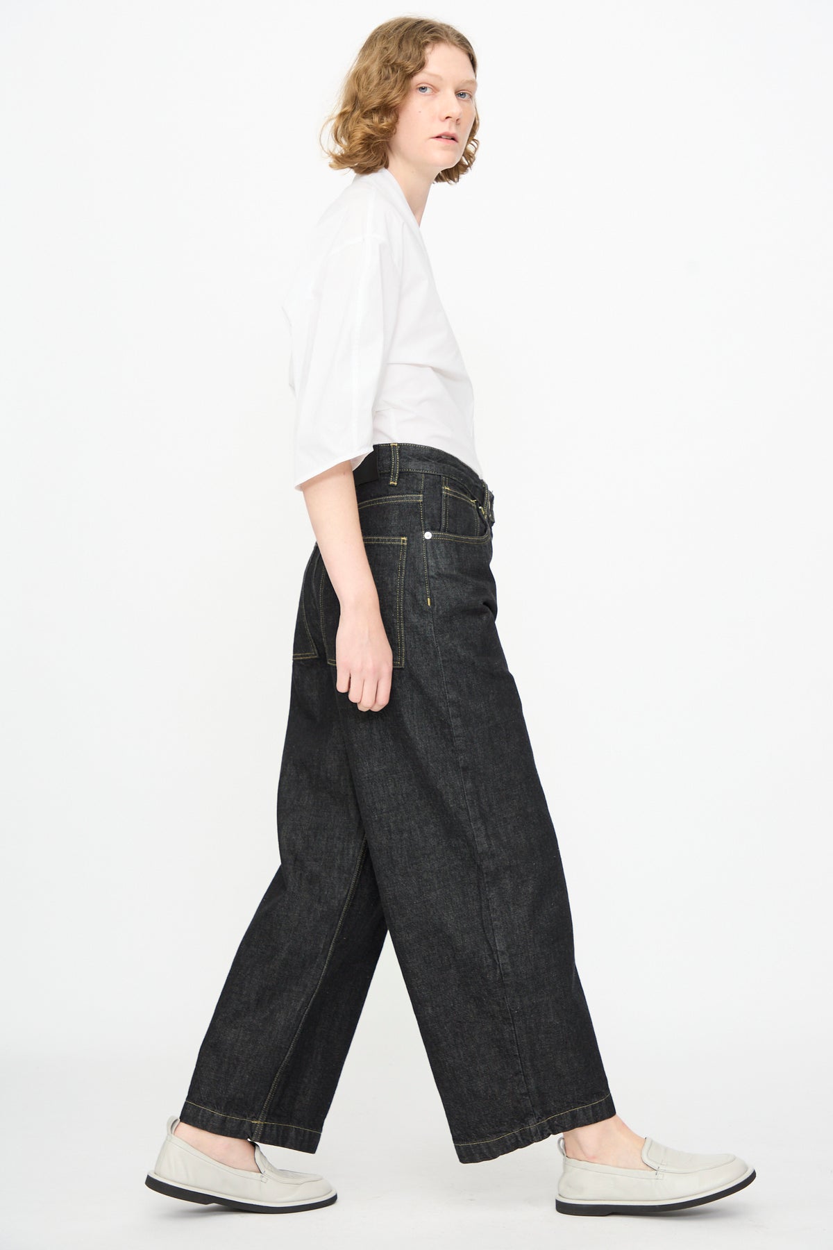 A person with short, wavy hair wears a white shirt, Studio Nicholson’s 13 oz. Denim Nieve Pant in Black—wide-leg and crafted from Italian denim—and white shoes, standing sideways against a plain white background.