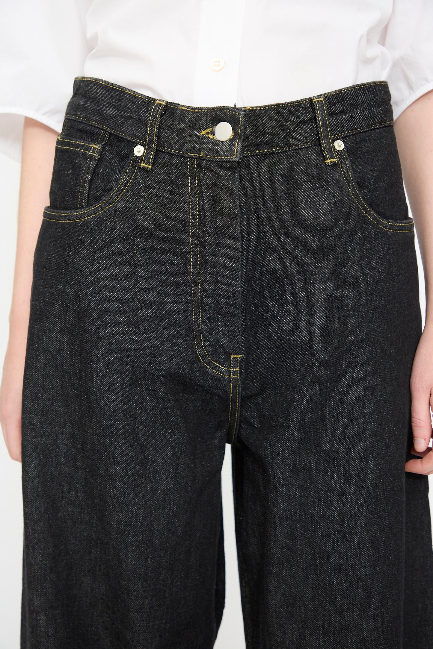 A person wearing Studio Nicholson’s 13 oz. Denim Nieve Pant in Black, crafted from luxurious Italian denim with yellow stitching, paired with a white button-up shirt.