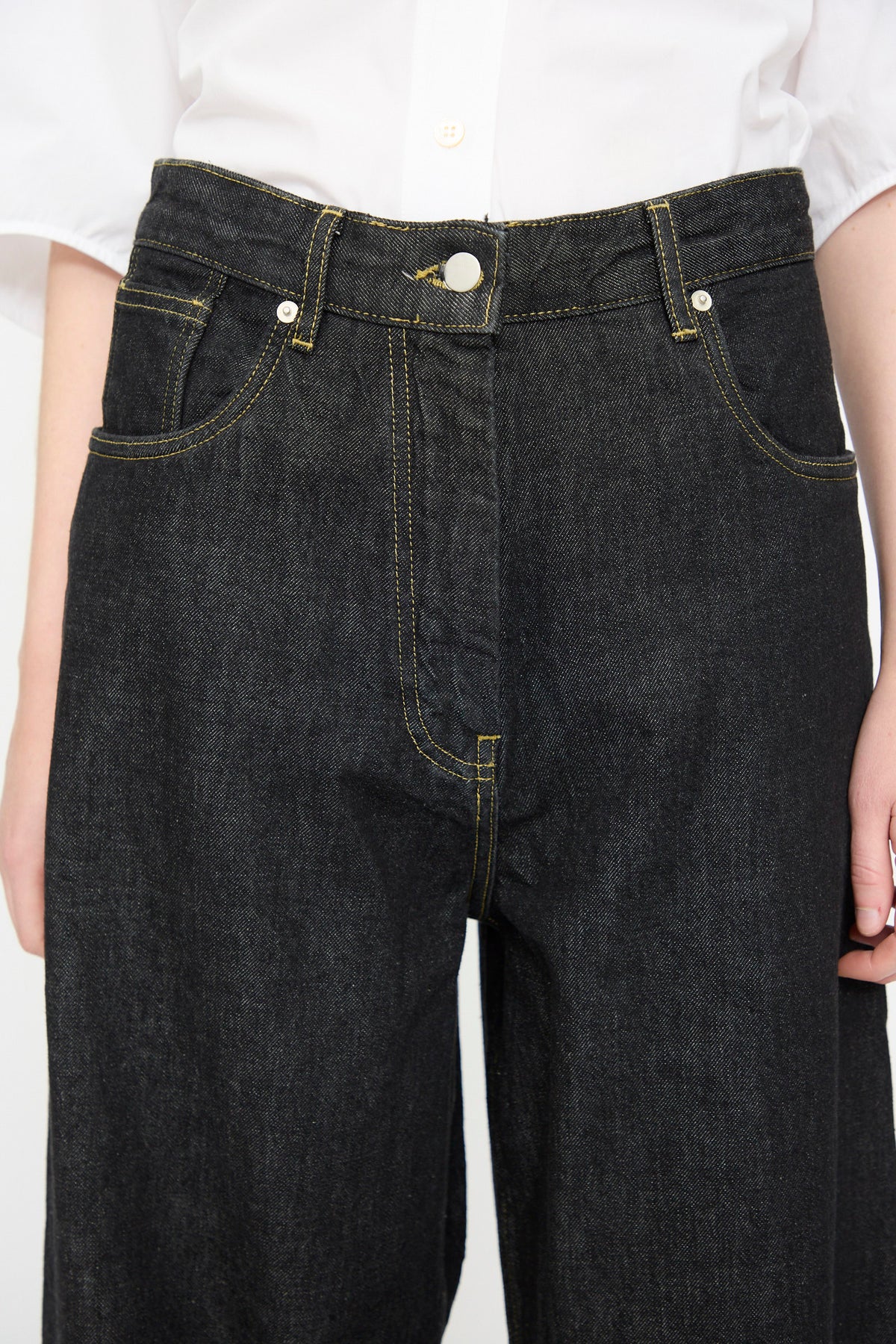 A person wearing Studio Nicholson’s 13 oz. Denim Nieve Pant in Black, crafted from luxurious Italian denim with yellow stitching, paired with a white button-up shirt.