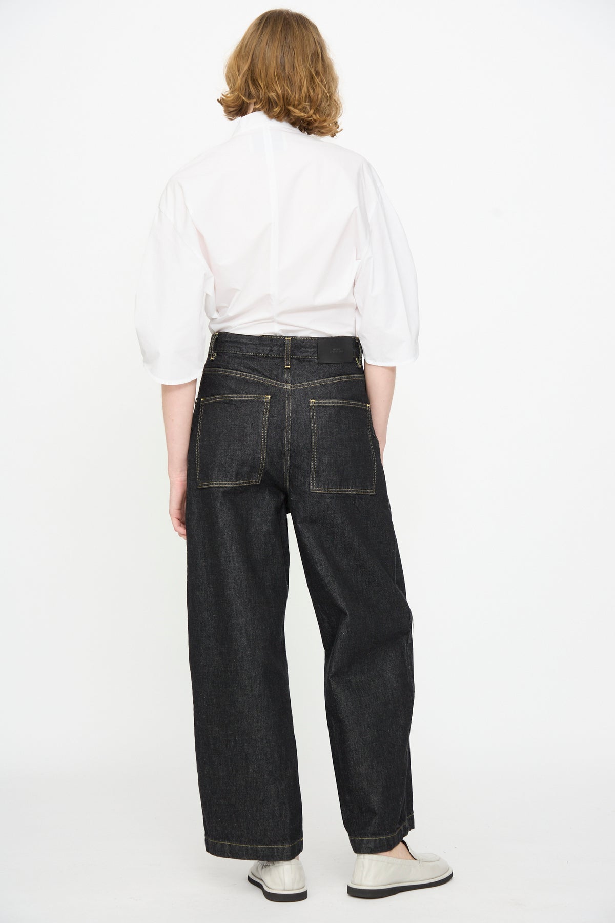 A person stands facing away against a plain white backdrop, wearing a white shirt with puffed sleeves, white shoes, and Studio Nicholson’s 13 oz. Denim Nieve Pant in Black—relaxed fit, wide-leg jeans crafted from premium Italian denim with back pockets.