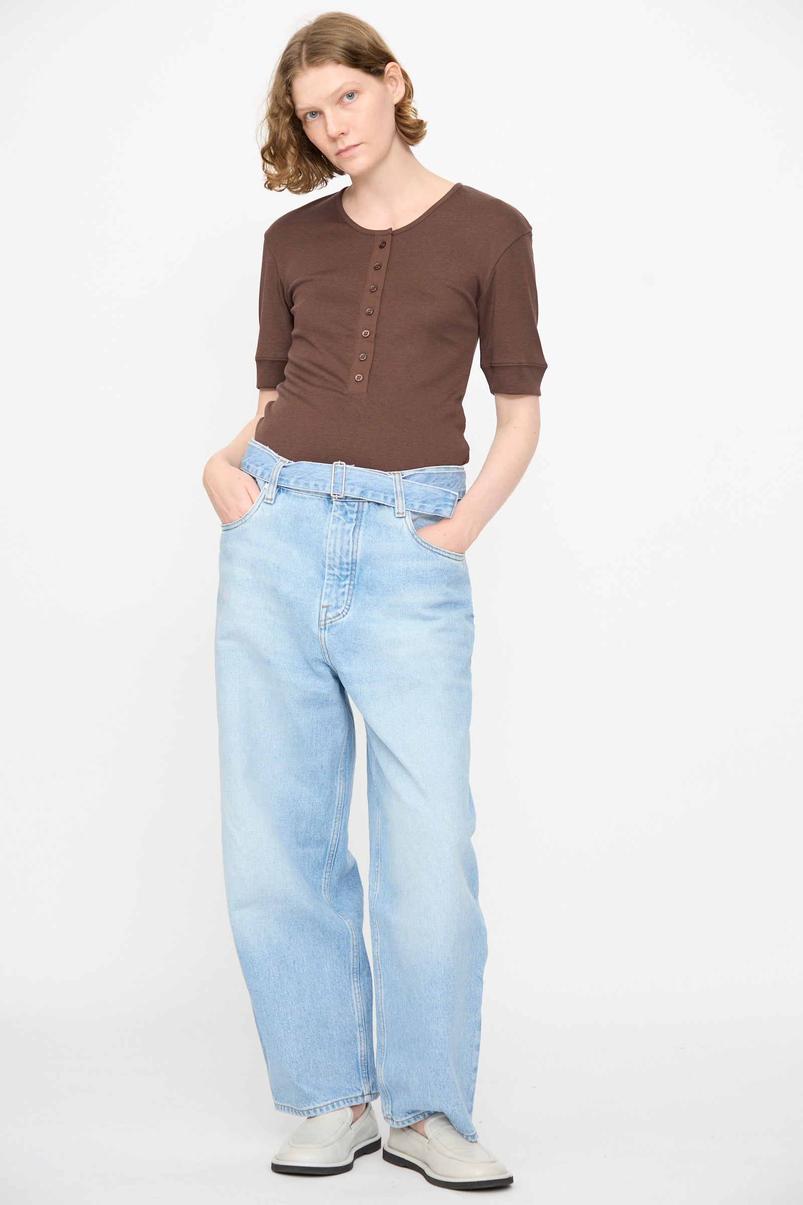 A person stands against a white background, wearing a brown short-sleeve buttoned top with a belted waist, white shoes, and Studio Nicholson’s 14 oz. Denim Cahun Pant in Bleached Wash—a light blue wide-leg relaxed denim—hands in pockets.