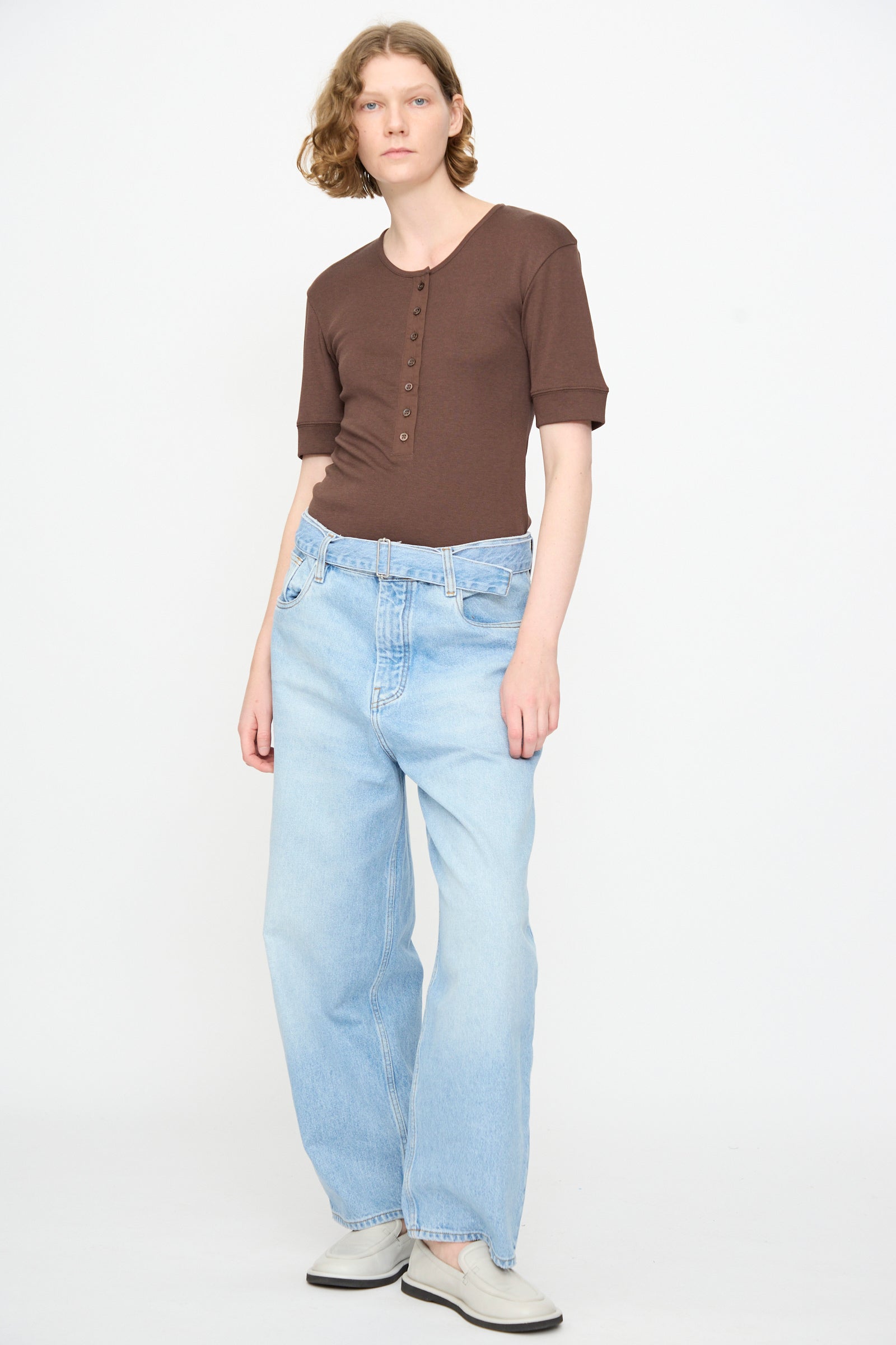 A person stands against a plain background, wearing a brown short-sleeve button-up shirt, Studio Nicholson 14 oz. Denim Cahun Pant in Bleached Wash with a belted waist, and white shoes.