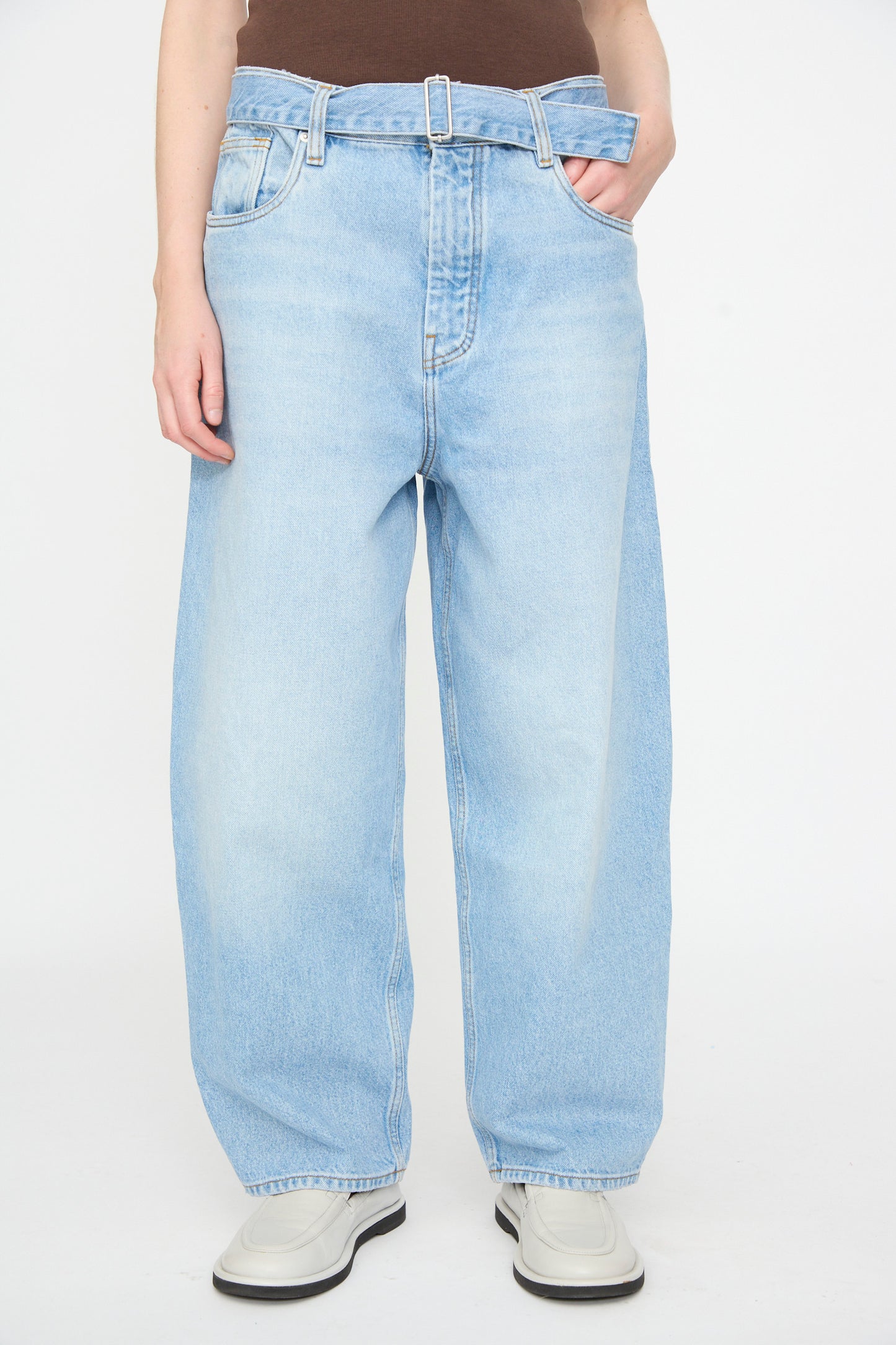 A person wears Studio Nicholson’s 14 oz. Denim Cahun Pant in Bleached Wash—featuring a baggy, relaxed silhouette with a folded waistband—paired with a brown top and light-colored shoes, standing against a plain white background.