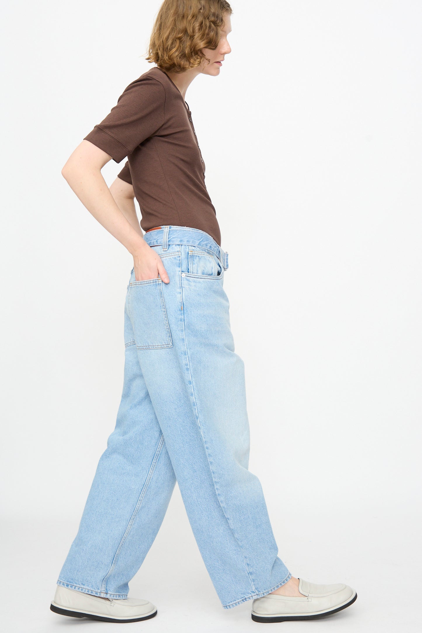 A person wearing a brown short-sleeve shirt, Studio Nicholson 14 oz. Denim Cahun Pant in Bleached Wash, and white shoes stands in profile against a plain white background.