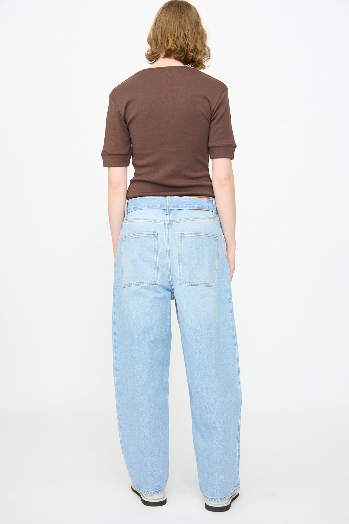A person faces away from the camera, wearing a short-sleeve brown shirt and Studio Nicholson 14 oz. Denim Cahun Pant in Bleached Wash—light blue, wide-leg relaxed jeans—while standing against a plain white background.