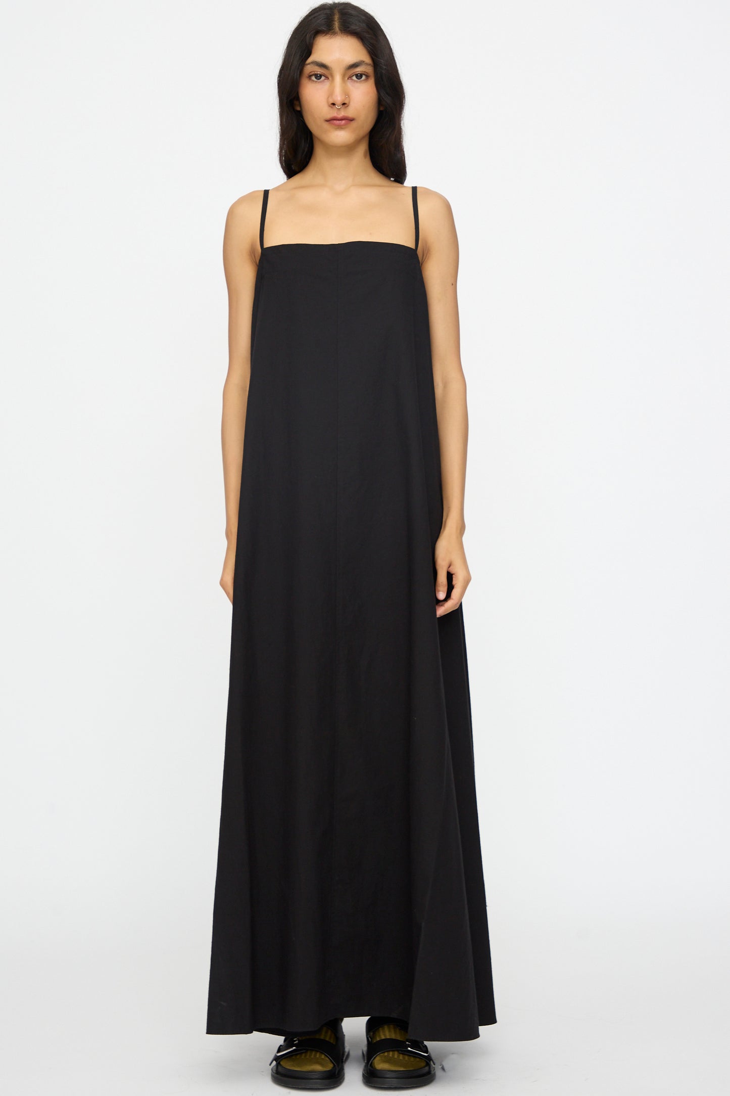 A woman faces forward against a plain white background, wearing the Studio Nicholson Achille Gathered Dress in Black— a sleeveless cotton maxi with thin straps and square neckline — paired with black shoes.