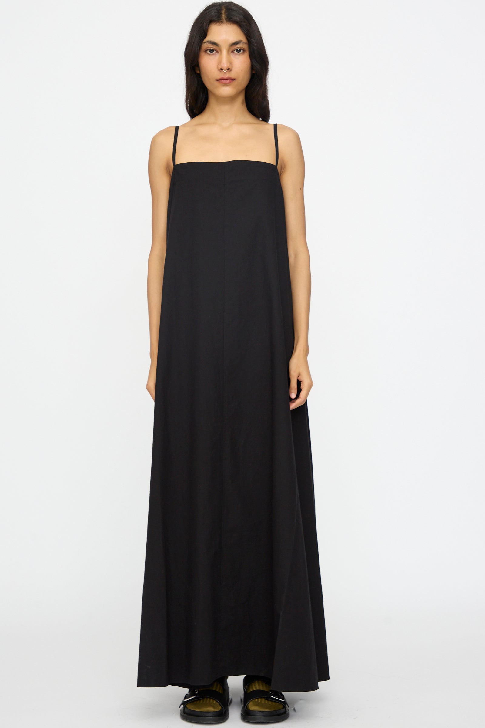 A woman faces forward against a plain white background, wearing the Studio Nicholson Achille Gathered Dress in Black— a sleeveless cotton maxi with thin straps and square neckline — paired with black shoes.