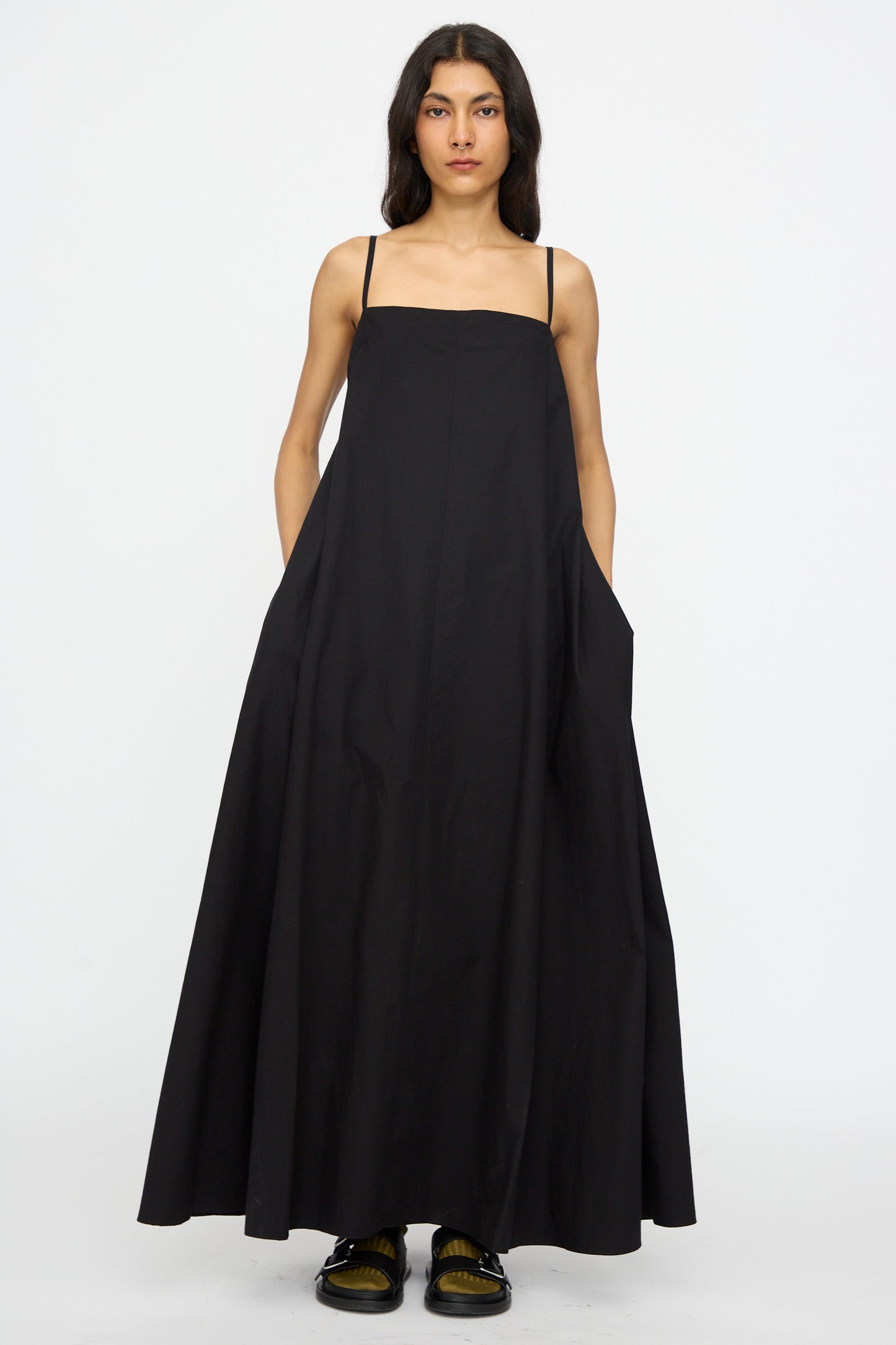 A person faces forward wearing the Studio Nicholson Achille Gathered Dress in Black—a cotton maxi dress with thin straps, side pockets, and a square neckline—paired with black sandals against a plain white background.