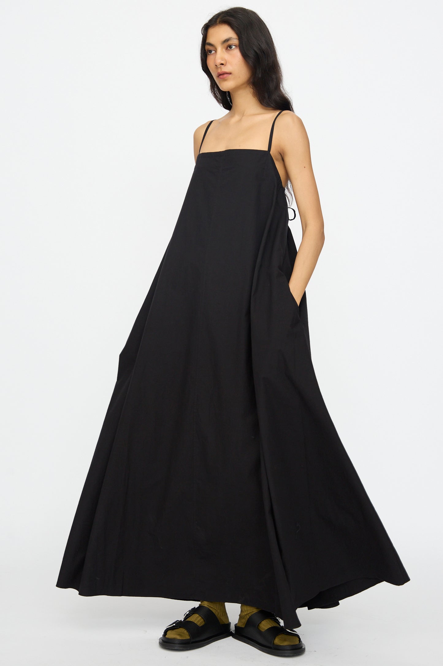 A person stands against a plain white background wearing the Studio Nicholson Achille Gathered Dress in Black, a loose-fitting cotton maxi dress with spaghetti straps and a square neckline, paired with yellow socks and black sandals.