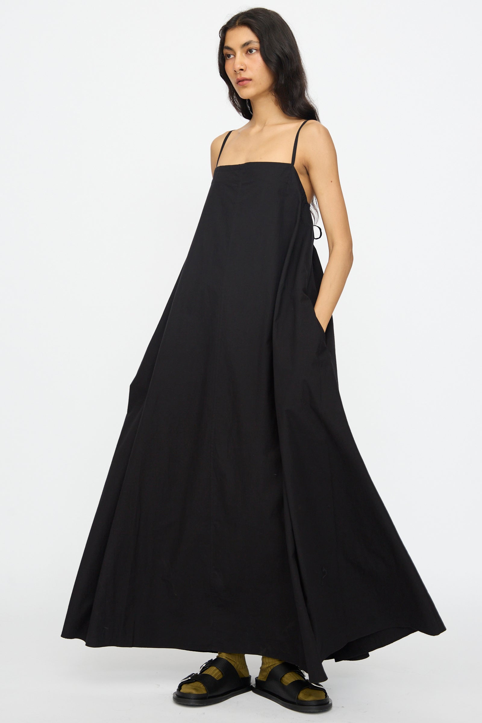 A person stands against a plain white background wearing the Studio Nicholson Achille Gathered Dress in Black, a loose-fitting cotton maxi dress with spaghetti straps and a square neckline, paired with yellow socks and black sandals.